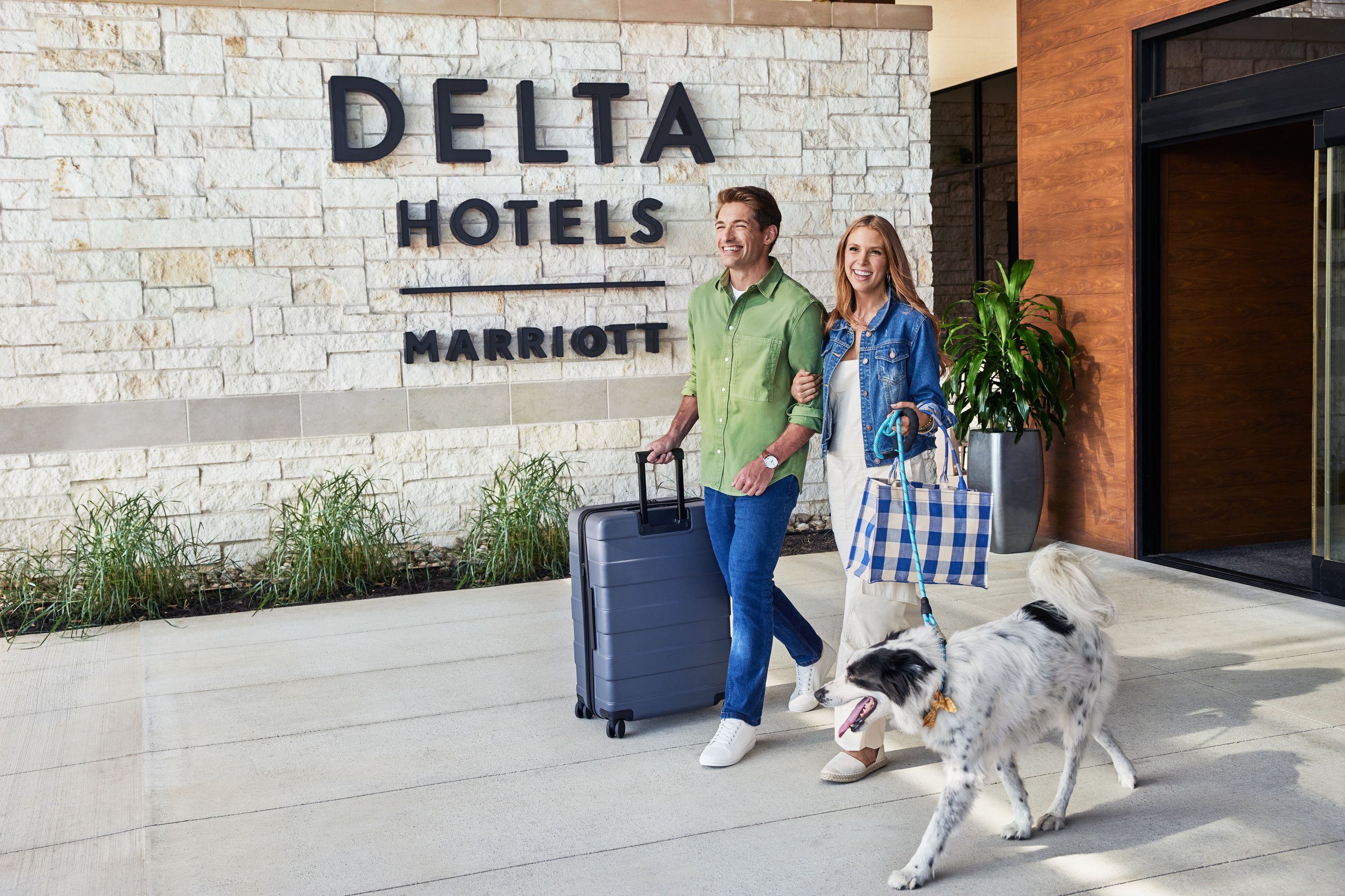 A smiling man and woman walk with a dog outside the Delta Hotels by Marriott entrance, with the man pulling a suitcase.