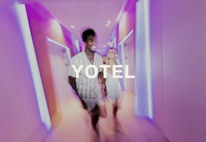 Two young people walking down a brightly lit hotel corridor with purple and pink neon lighting, smiling and enjoying their stay.