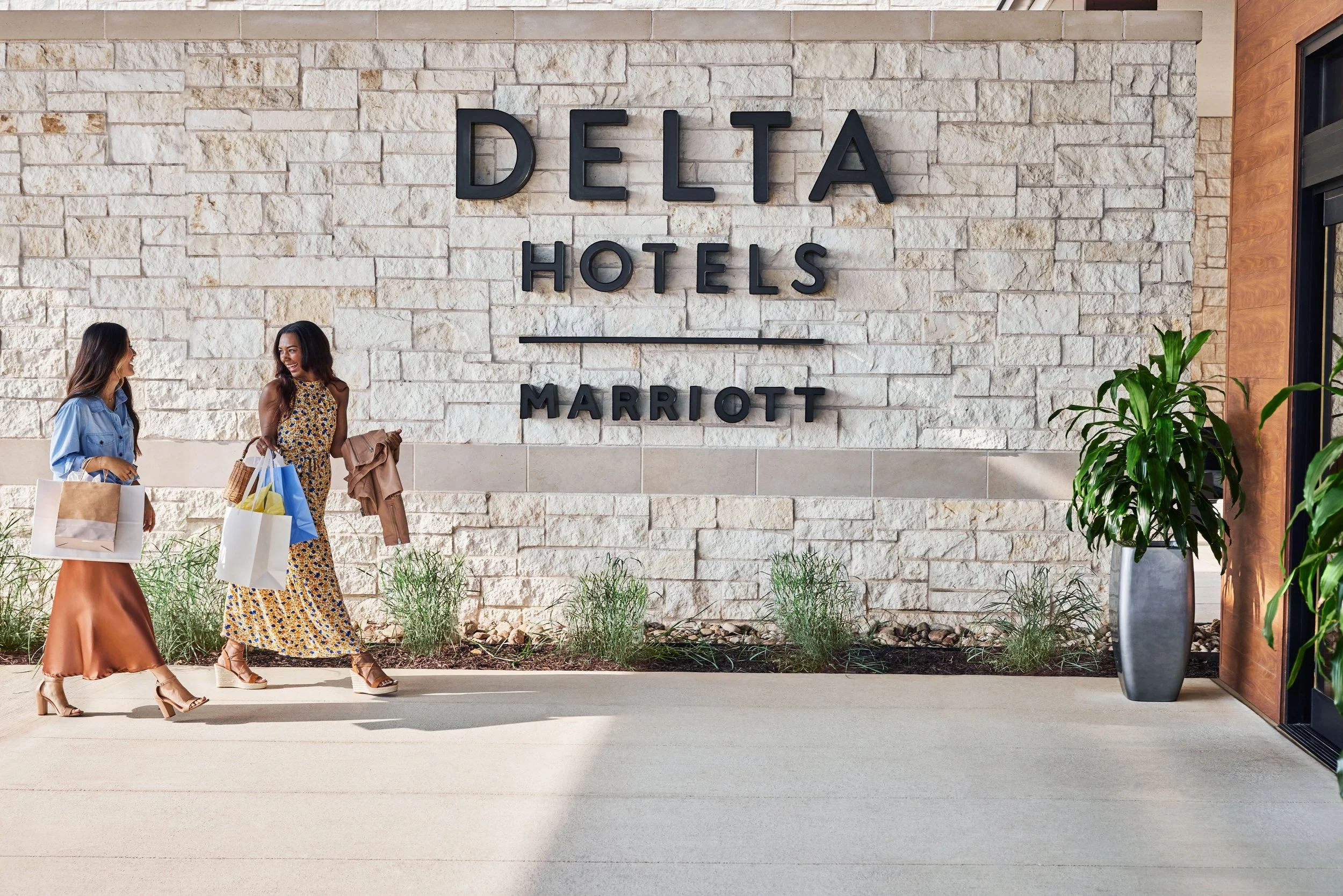 Two women walking past the entrance of a hotel with a large sign that reads 'Delta Hotels Marriott' on a stone wall.