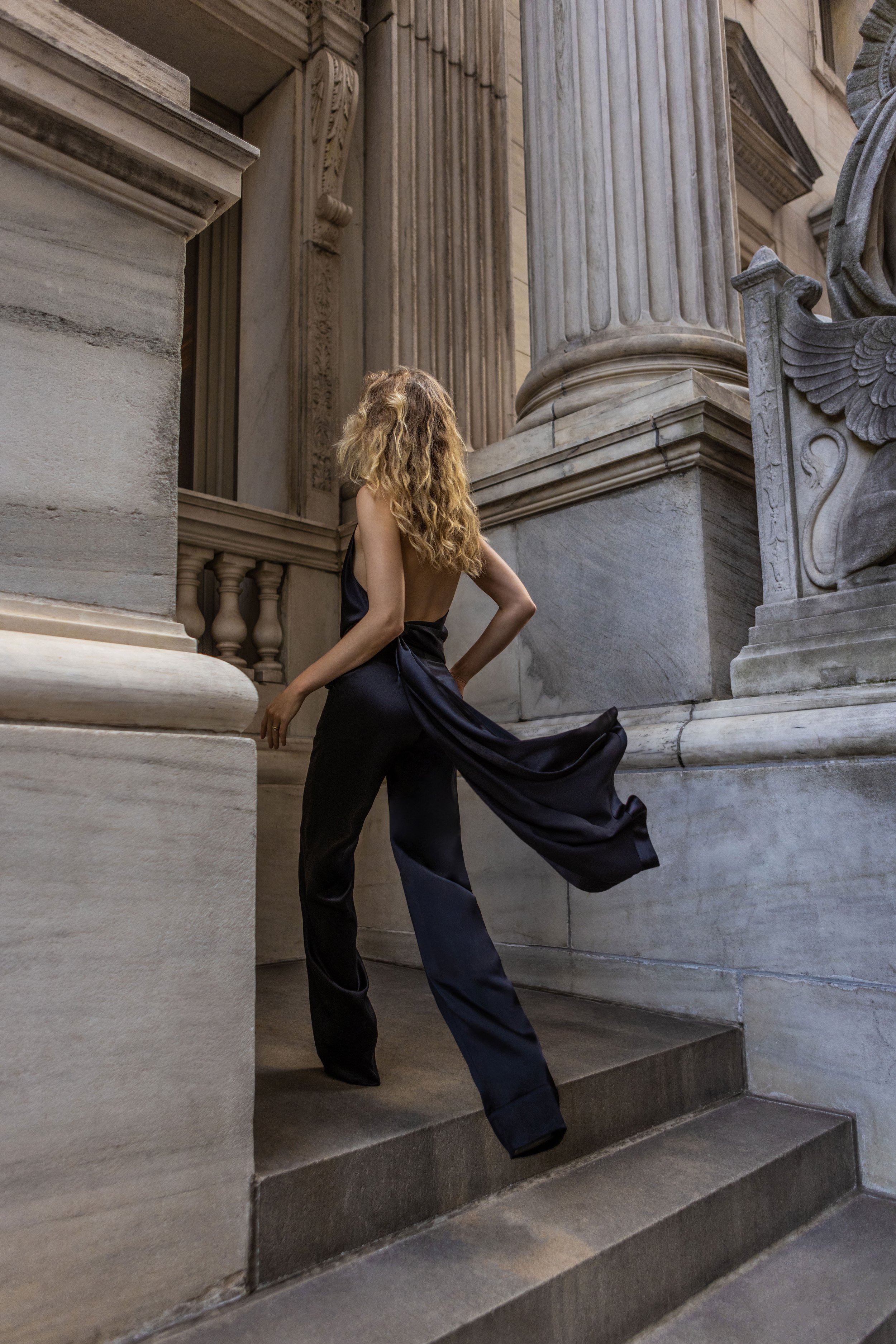 A woman in a black satin jumpsuit standing on stone steps inside a building with classical architecture, surrounded by large marble columns and ornate details.