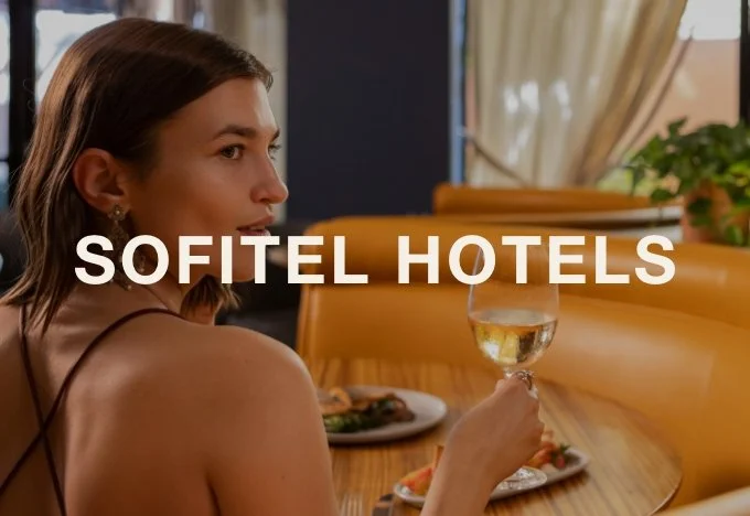 A woman with brown hair sitting at a restaurant table holding a glass of white wine, with the words 'Sofitel Hotels' overlaid on the image.