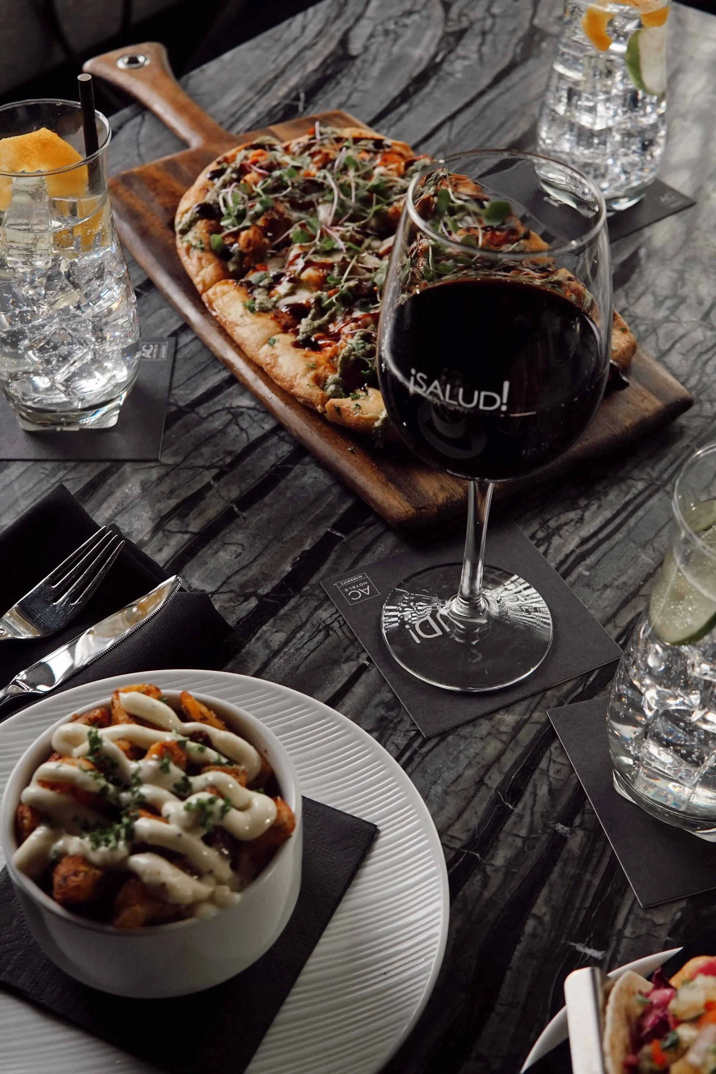 A table set with a slice of pizza on a wooden platter, a bowl of loaded fries with sauce and herbs, a glass of red wine, and bottles of water with lime and lemon slices.