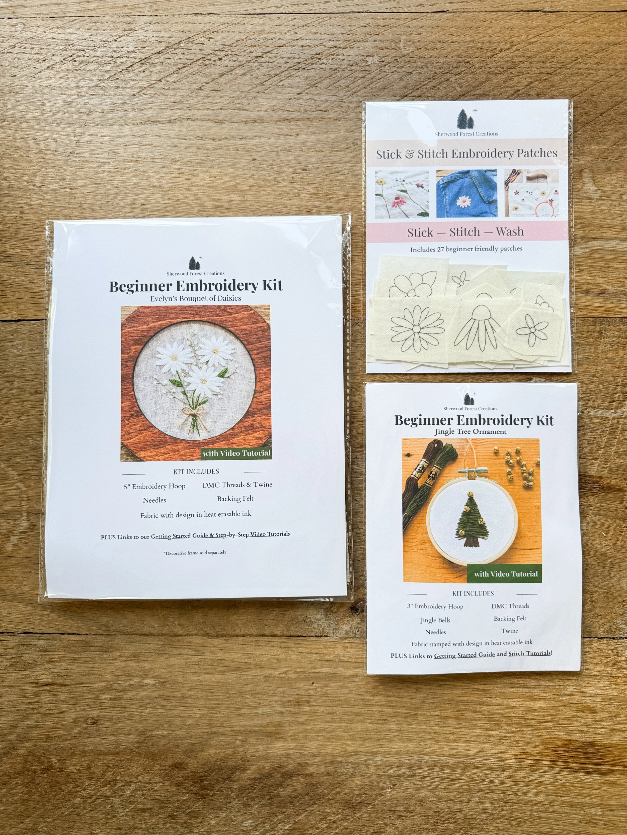 Two embroidery kit packages on a wooden surface, one for beginners with a daisy design and the other with a Christmas tree design, both with instructional videos.