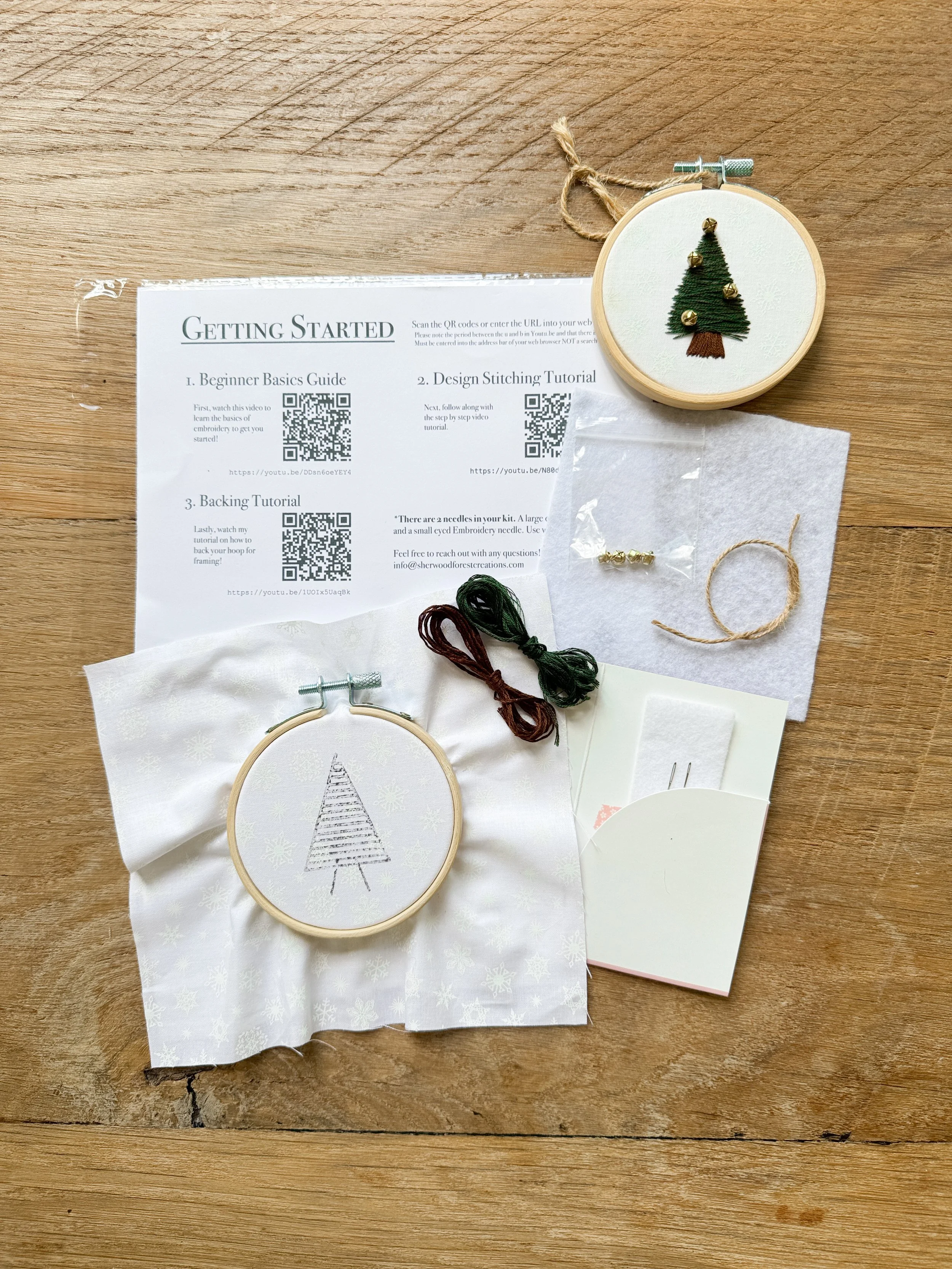 Embroidery hoop with a Christmas tree design on white fabric, surrounded by embroidery supplies and instruction sheets on a wooden surface.