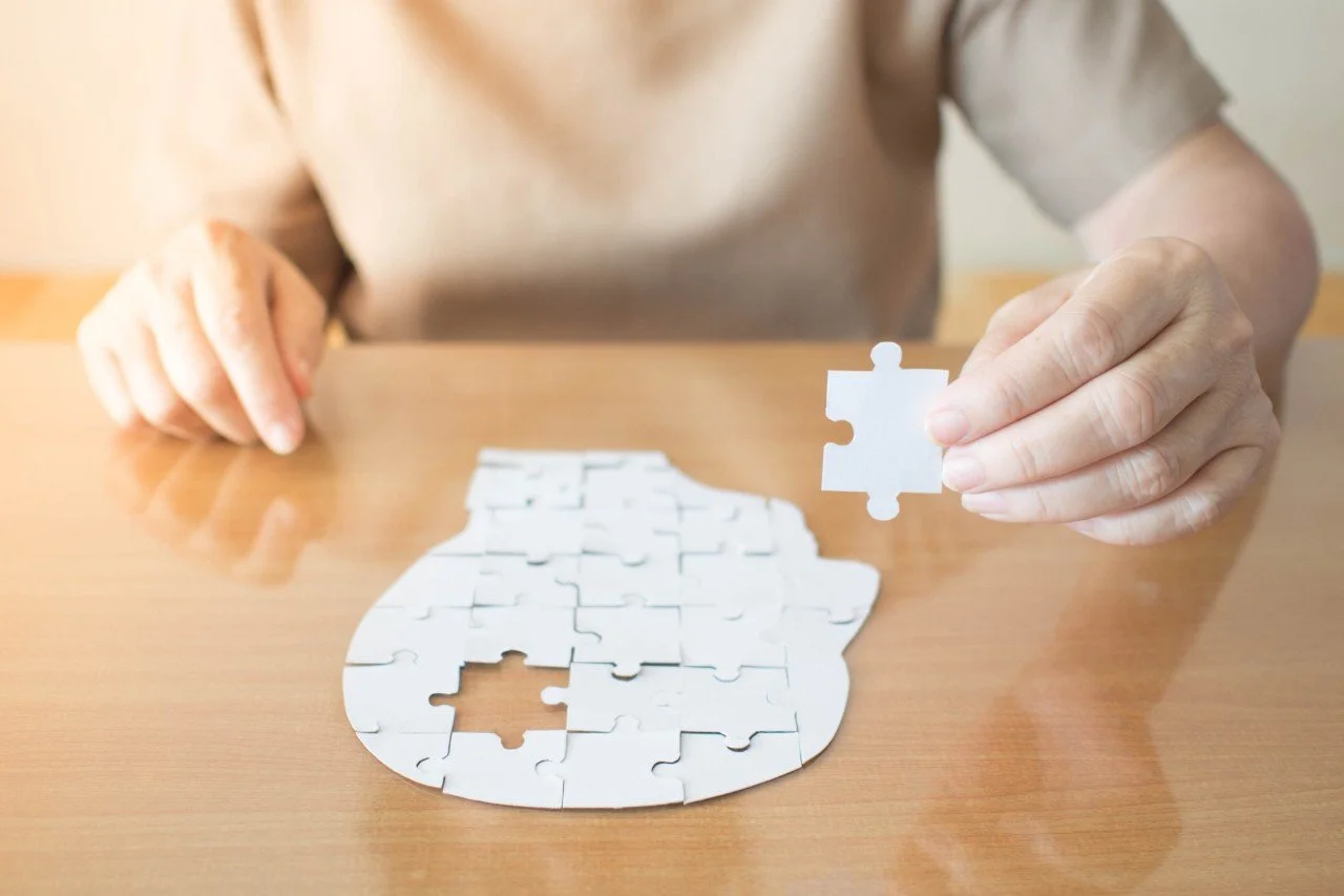 Person assembling a white jigsaw puzzle on a wooden table, holding a puzzle piece in their hand