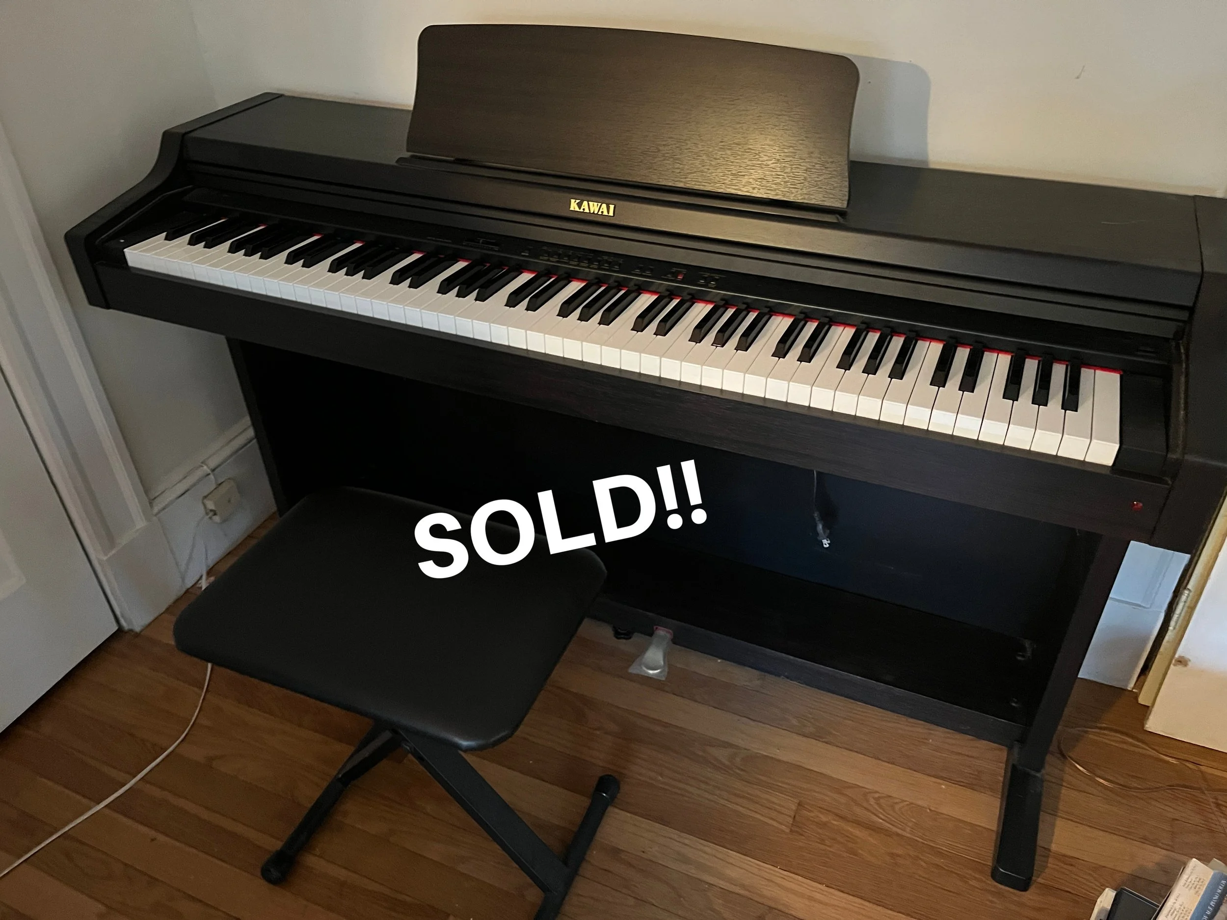 A black digital Kawai piano with a bench placed in front of it, and a "SOLD!!" sign over the bench.
