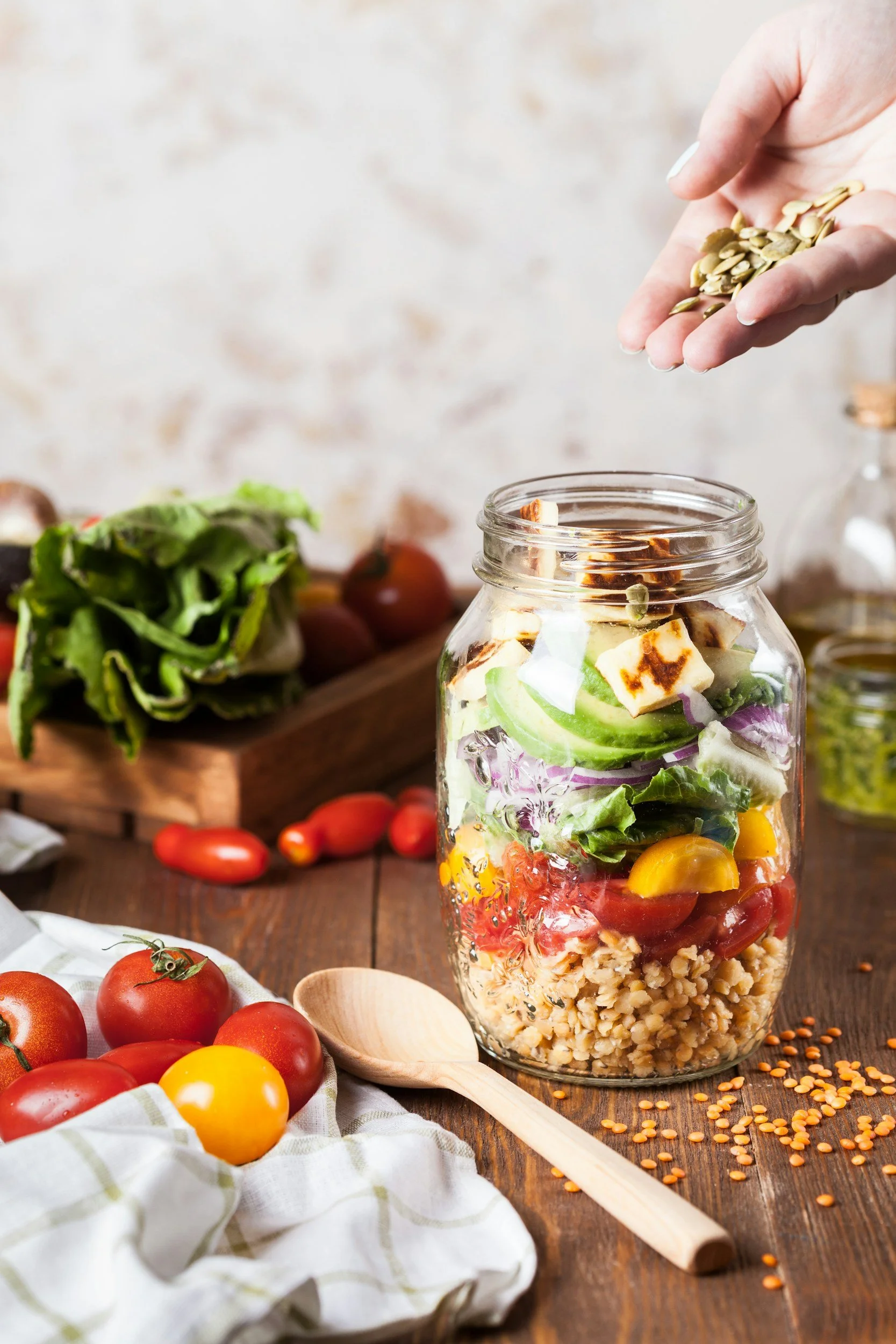Glass jar filled with layers of salad ingredients including cherry tomatoes, chopped cucumbers, red onions, tofu, and cooked lentils, on a rustic wooden table with scattered lentils, fresh tomatoes, and a striped cloth.