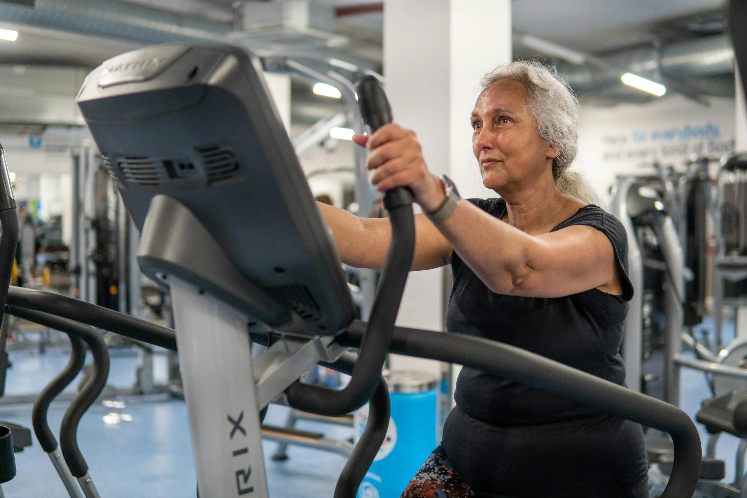 An elderly woman with gray hair working out on a treadmill in a gym, wearing a black shirt and patterned leggings.