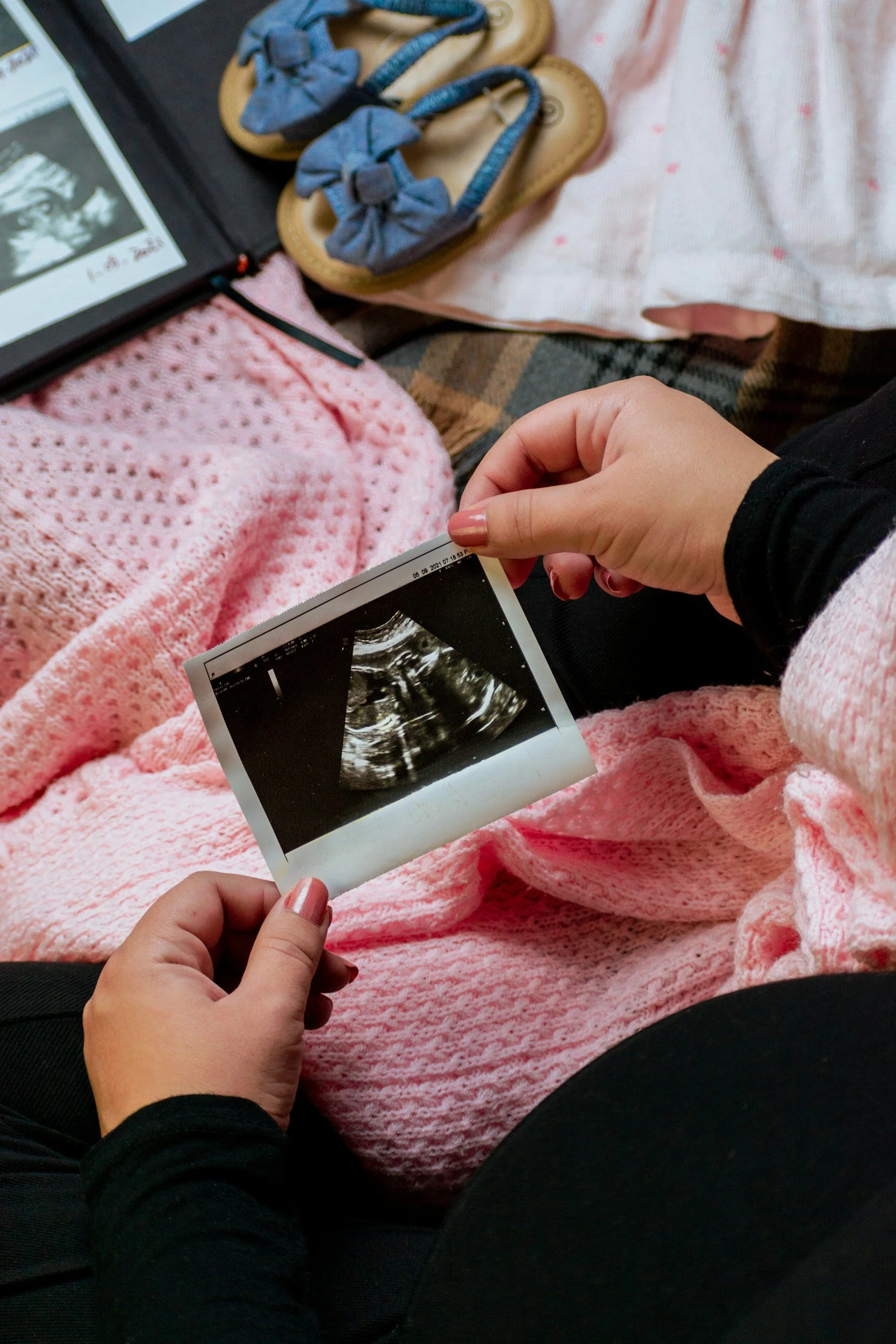 A person's hand holding an ultrasound scan image of a fetus. They are sitting with pink, knitted fabric, and other personal items are nearby, including blue sandals, a blanket, and a smartphone or tablet.