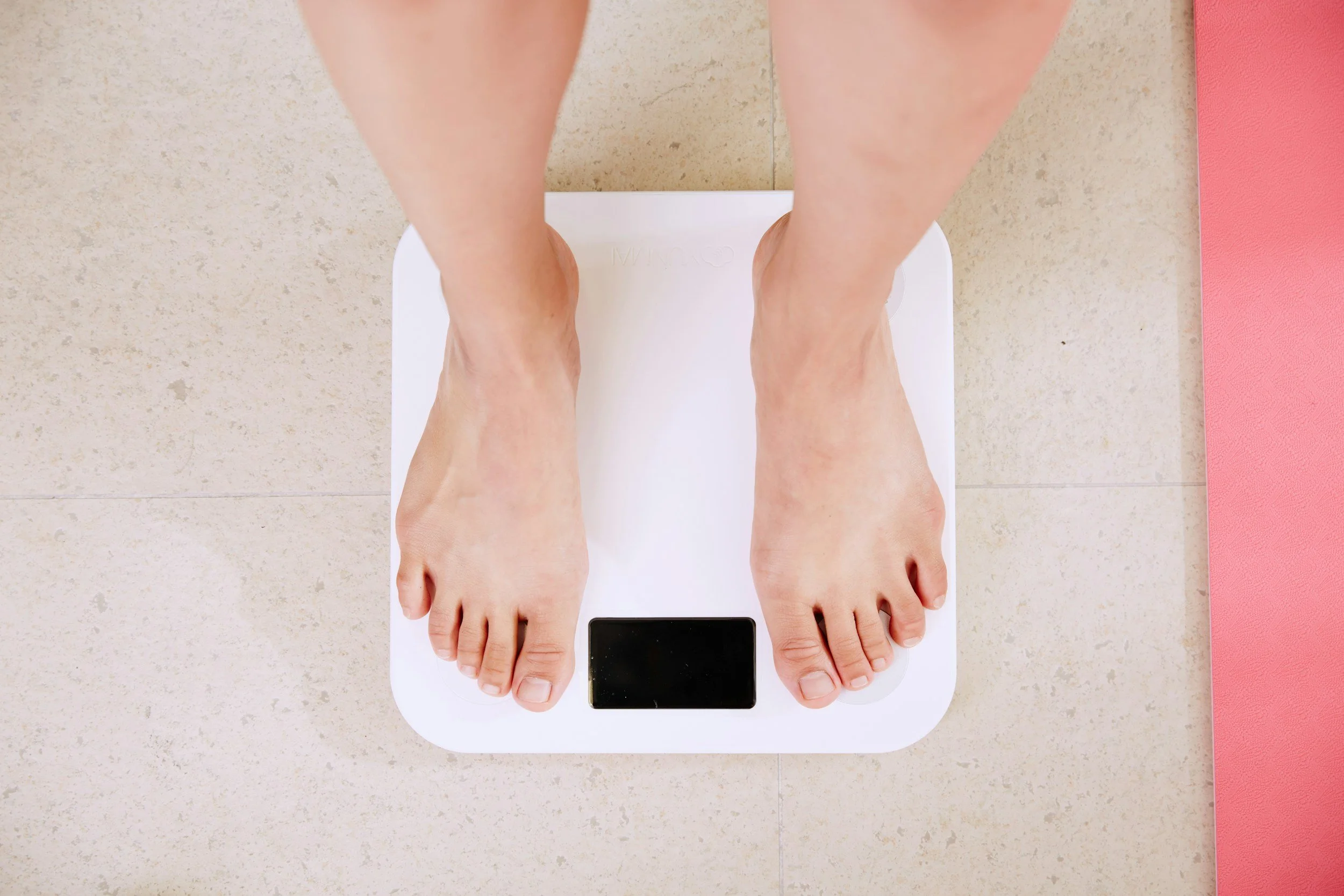 Overhead view of a person standing on a digital bathroom scale, with a pink yoga mat to the right on a beige tiled floor.