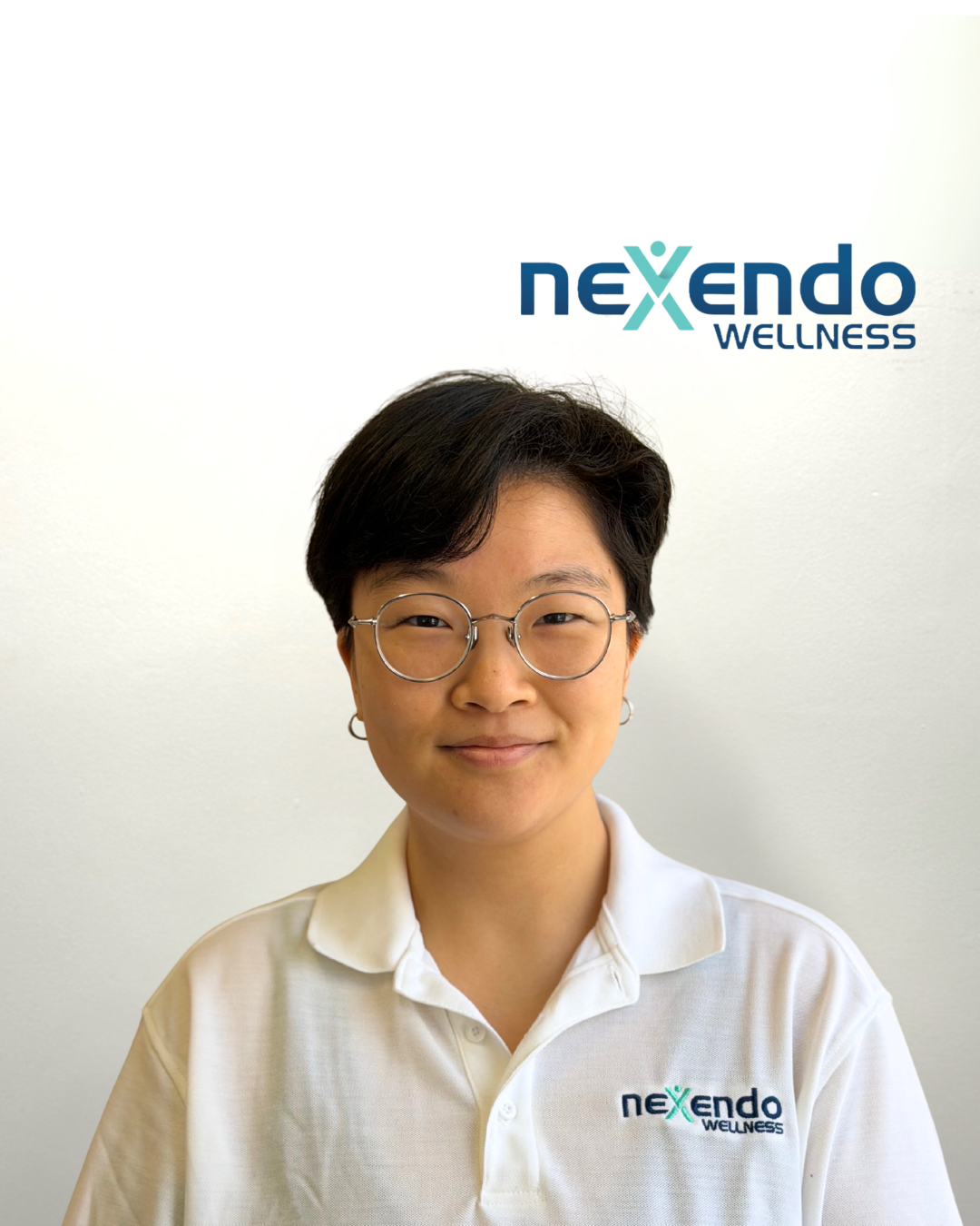 A woman with short dark hair, wearing glasses and earrings, smiling at the camera in front of a white wall with Nexendo Wellness logo.