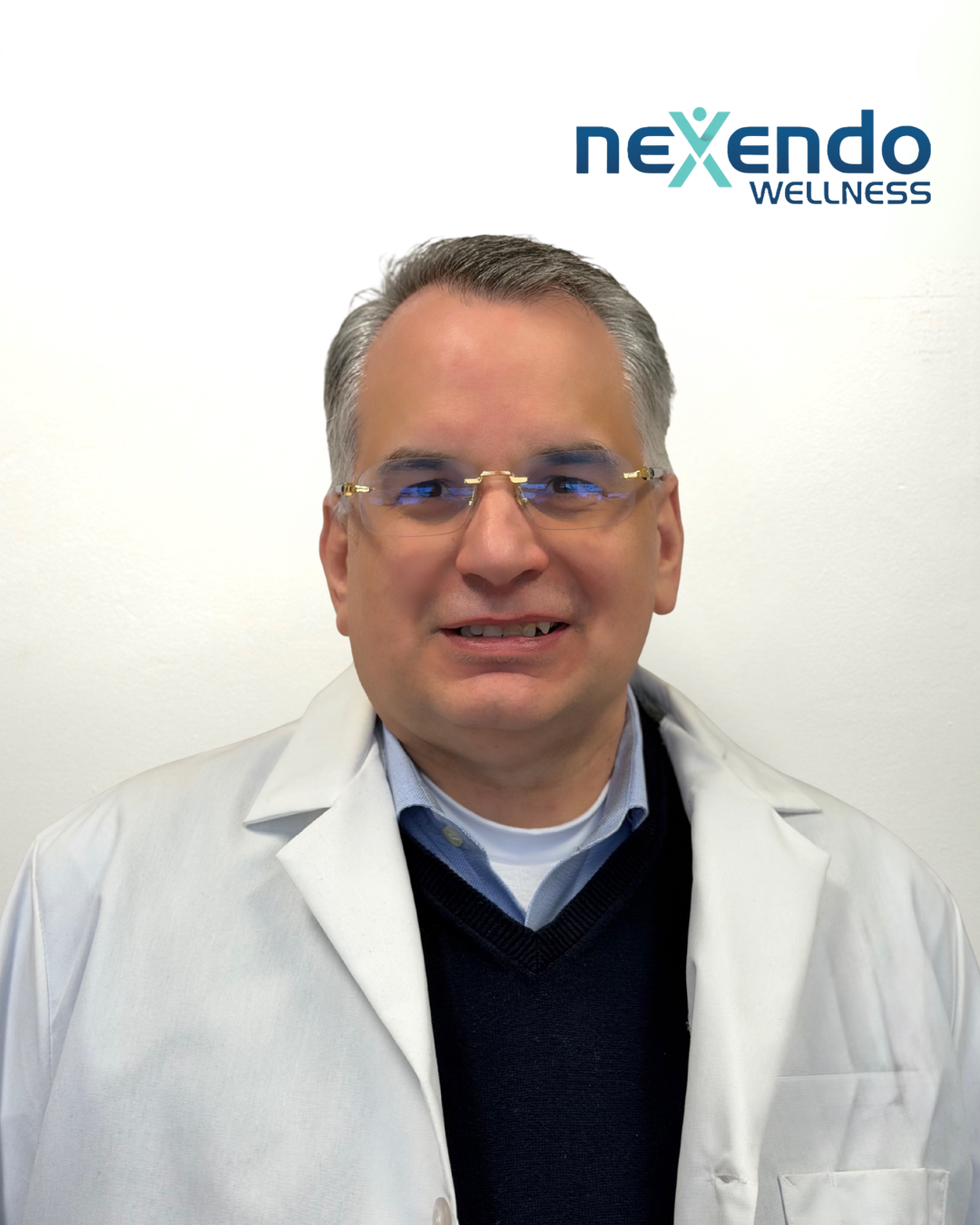 A man wearing glasses, a collared shirt, a dark sweater, and a white lab coat standing in front of a white wall with a Nexendo Wellness logo.