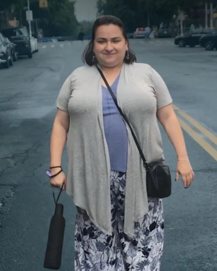 A woman walking on a city street during daytime, holding a black umbrella and wearing a gray cardigan, blue shirt, patterned pants, and carrying a small black purse.