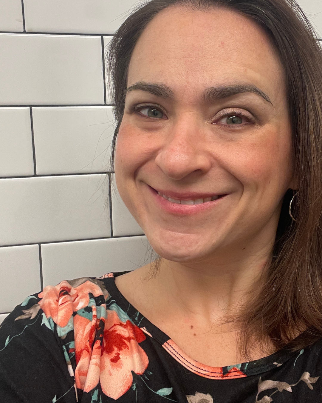A woman with light skin, brown hair, and blue eyes smiling in front of white subway tiles, wearing a black top with a pink and orange floral print and hoop earrings.
