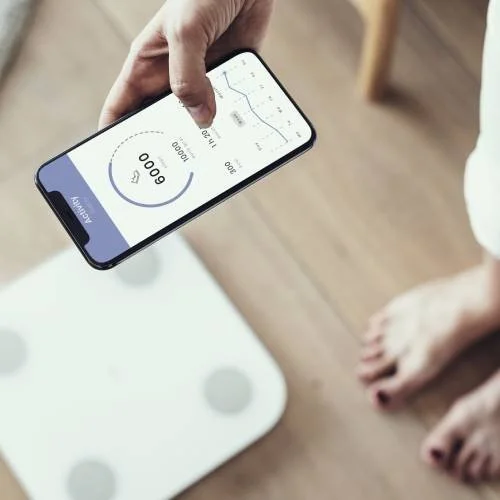 A person holding a smartphone showing a fitness app with a step count of 0 and a line graph, standing on a digital bathroom scale.