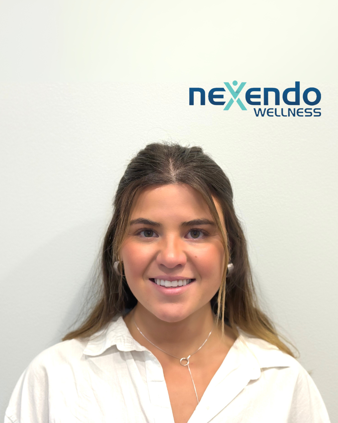 A woman smiling posing in front of a white wall with the Nexendo Wellness logo on it.