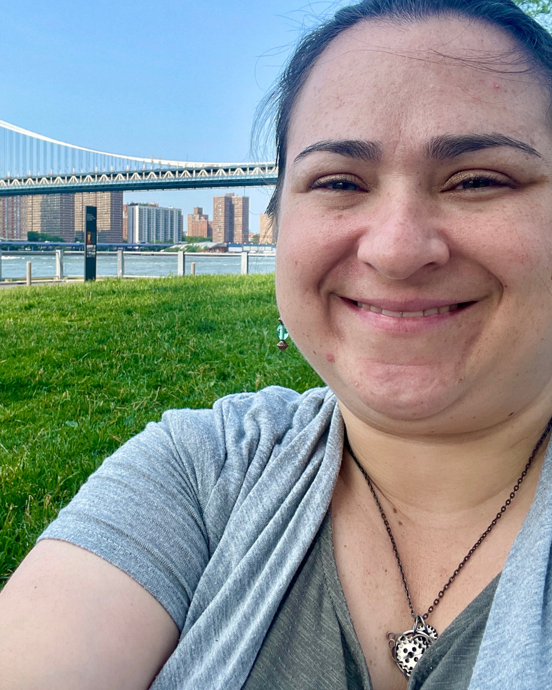 A woman smiling outdoors with a bridge and city skyline in the background, sitting on a grassy area near a body of water.
