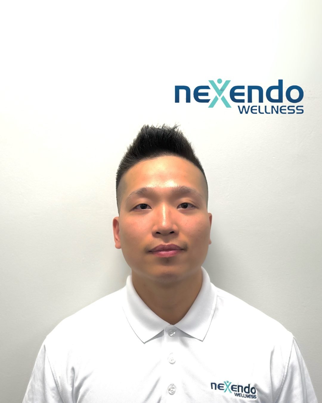 A man with short black hair wearing a white polo shirt with a 'Nexendo Wellness' logo, standing in front of a white wall with a 'Nexendo Wellness' logo.