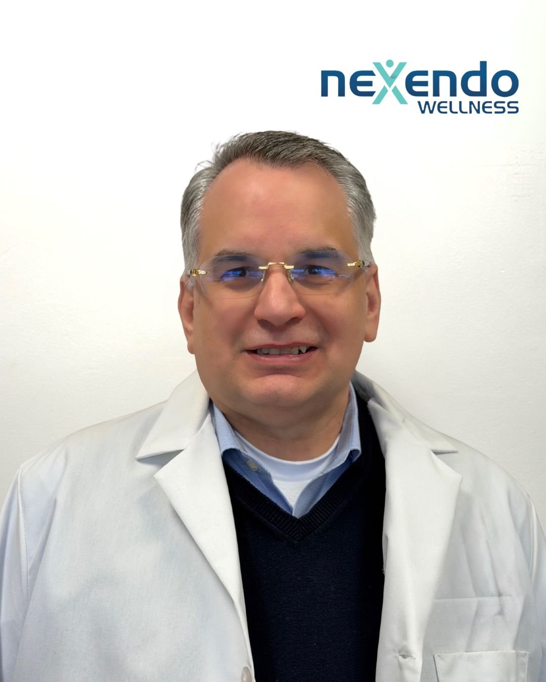 A man with gray hair and glasses wearing a white medical coat standing in front of a white wall with a Nexendo Wellness logo.