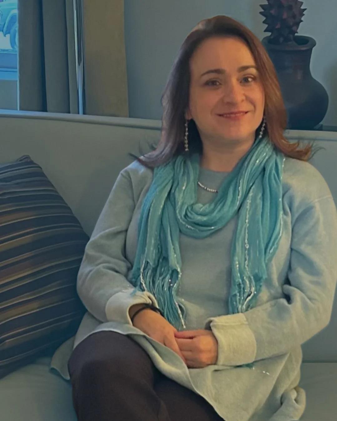 A woman sitting on a light-colored sofa, wearing a light gray sweatshirt, a turquoise scarf, and dark pants, smiling at the camera. There is a striped pillow beside her and a dark vase with a decorative object on top in the background.