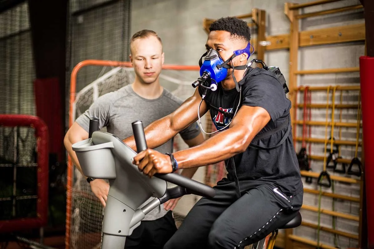 A man wearing a protective mask and exercising bike in a gym while another man supervises.
