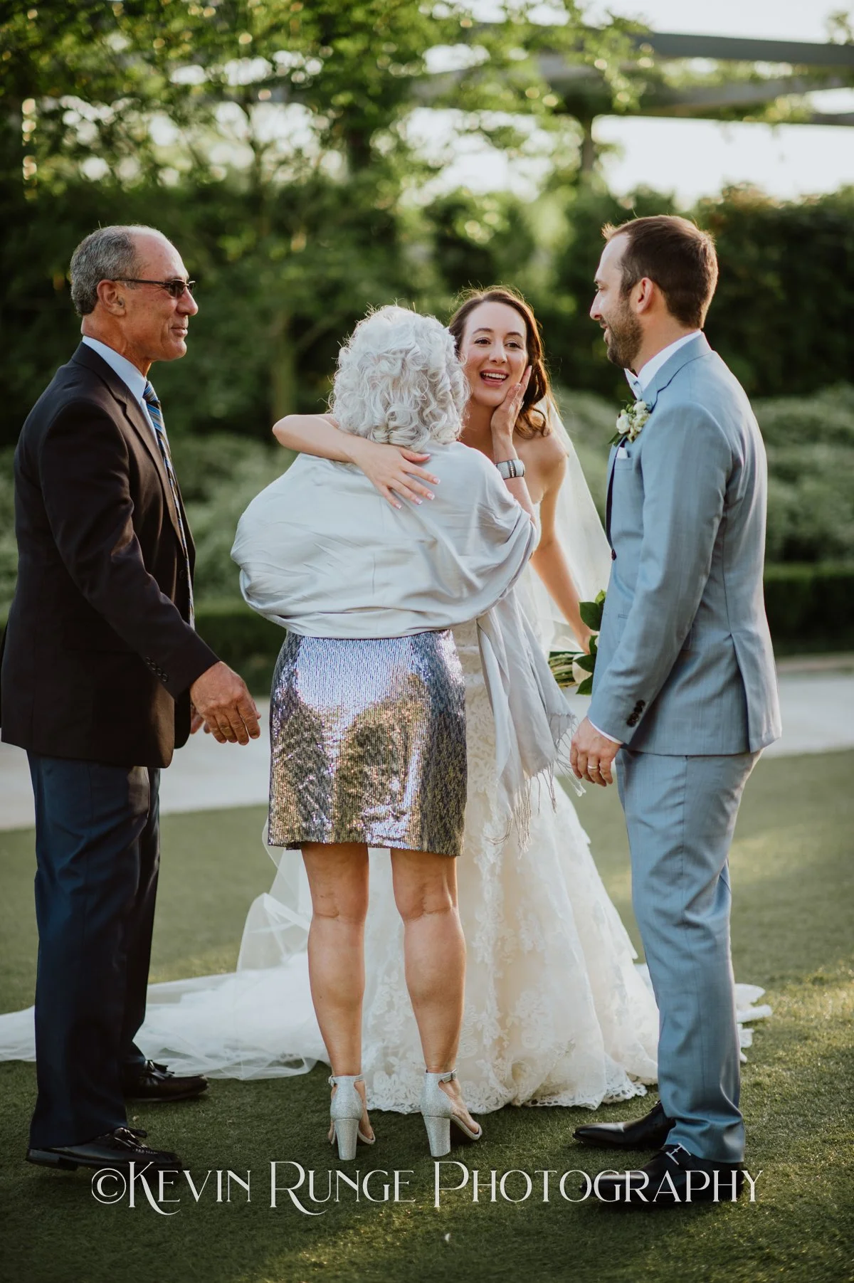 A wedding ceremony outdoors with a bride in a lace wedding dress, a groom in a light gray suit holding a bouquet, and three older adults, two women and one man, engaging emotionally, possibly parents or family members, on a lawn with trees in the bac