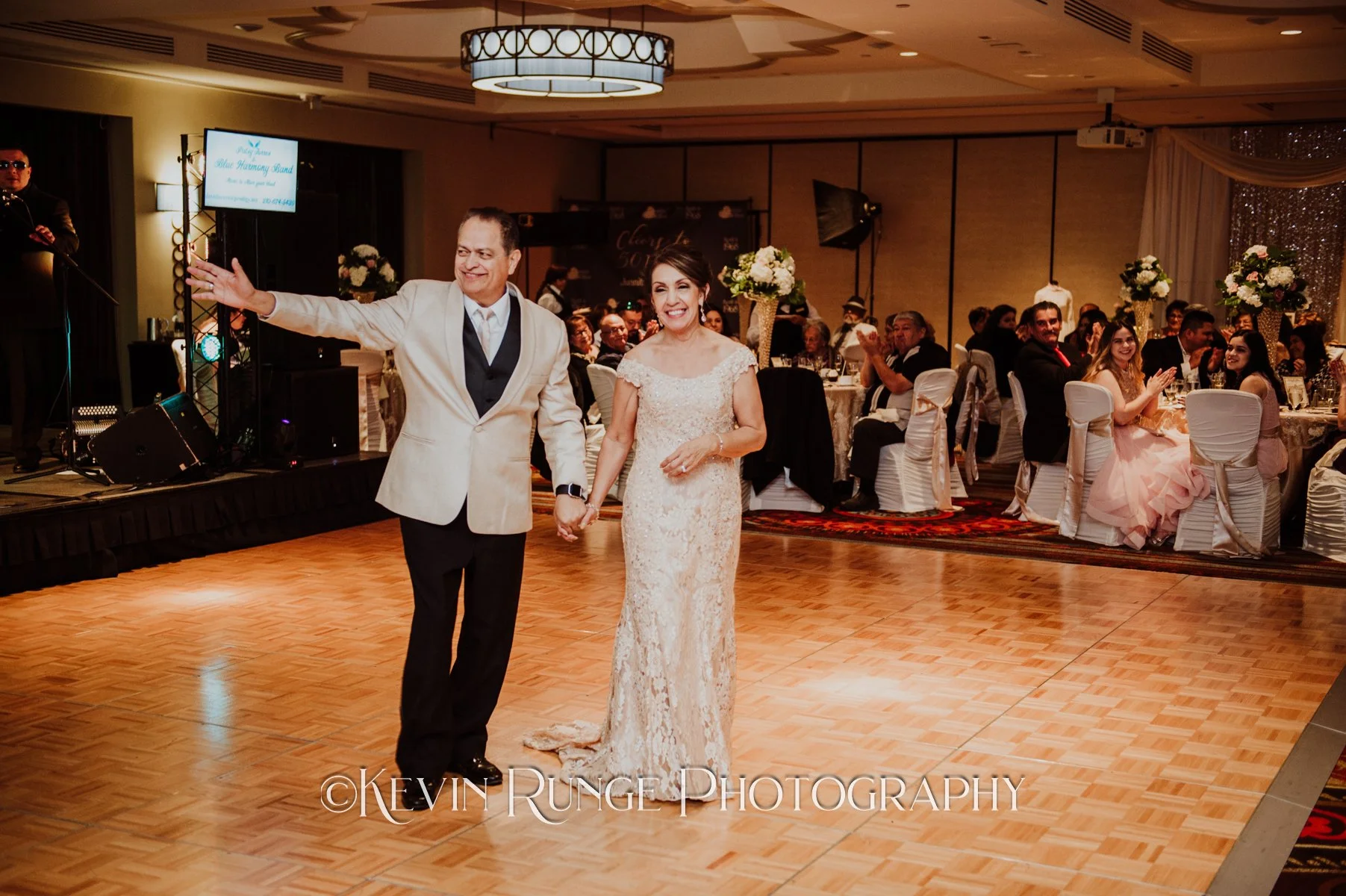 Bride and groom holding hands and smiling on the dance floor, with seated guests clapping and enjoying the celebration in the background at a wedding reception.