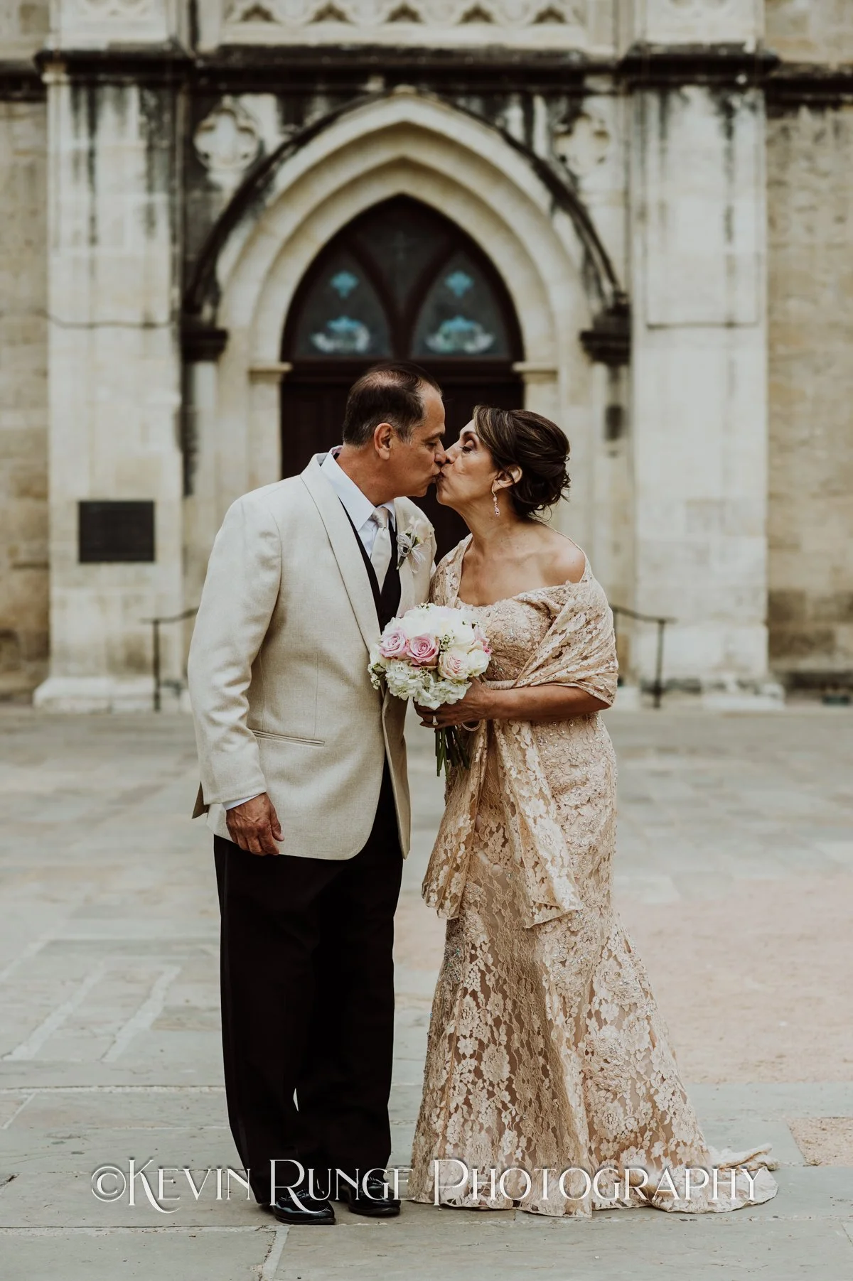 A couple dressed in wedding attire sharing a kiss outdoors, with a historic stone building in the background. The woman is holding a bouquet of pink and white flowers, and they are standing close together.
