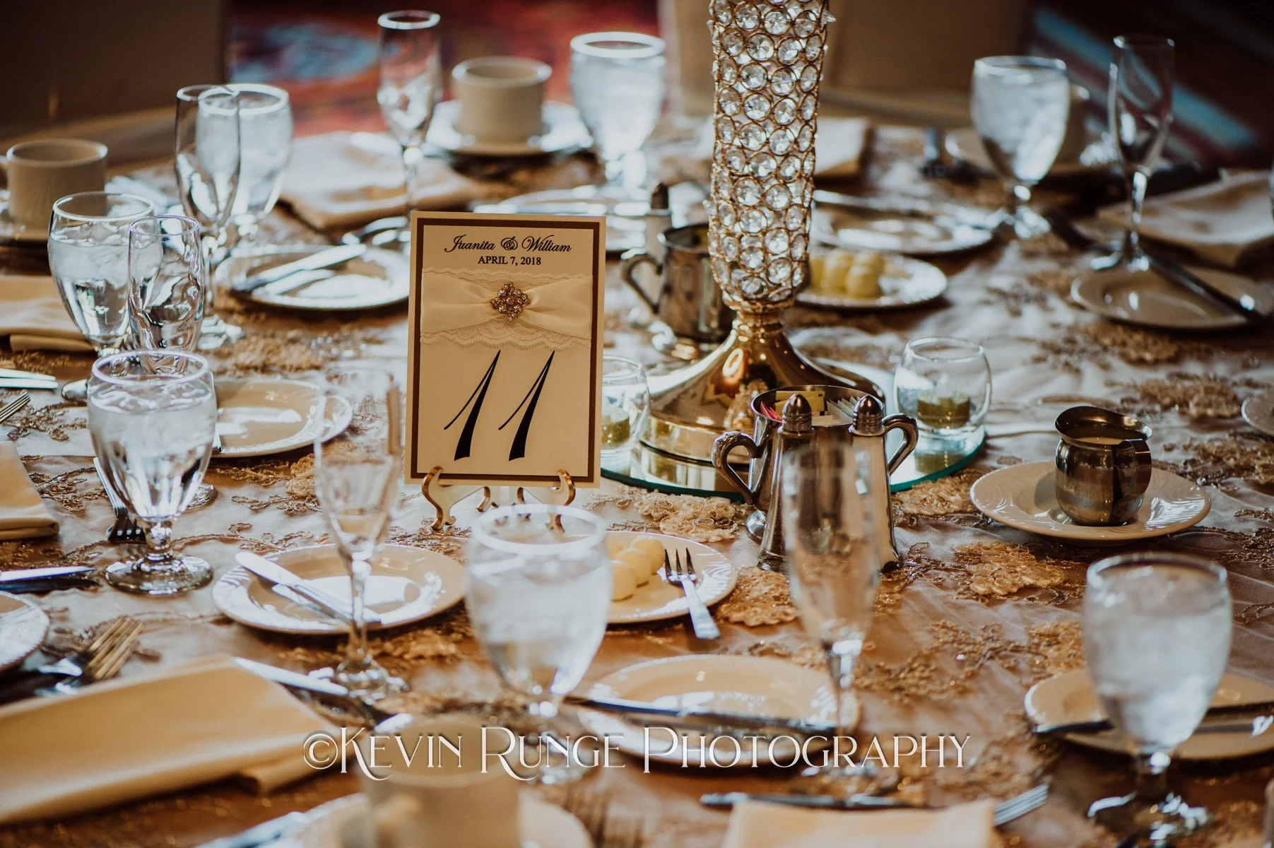 Elegant wedding reception table with glassware, plates, silverware, a table number card, a tall decorative centerpiece, and a cream-colored tablecloth.