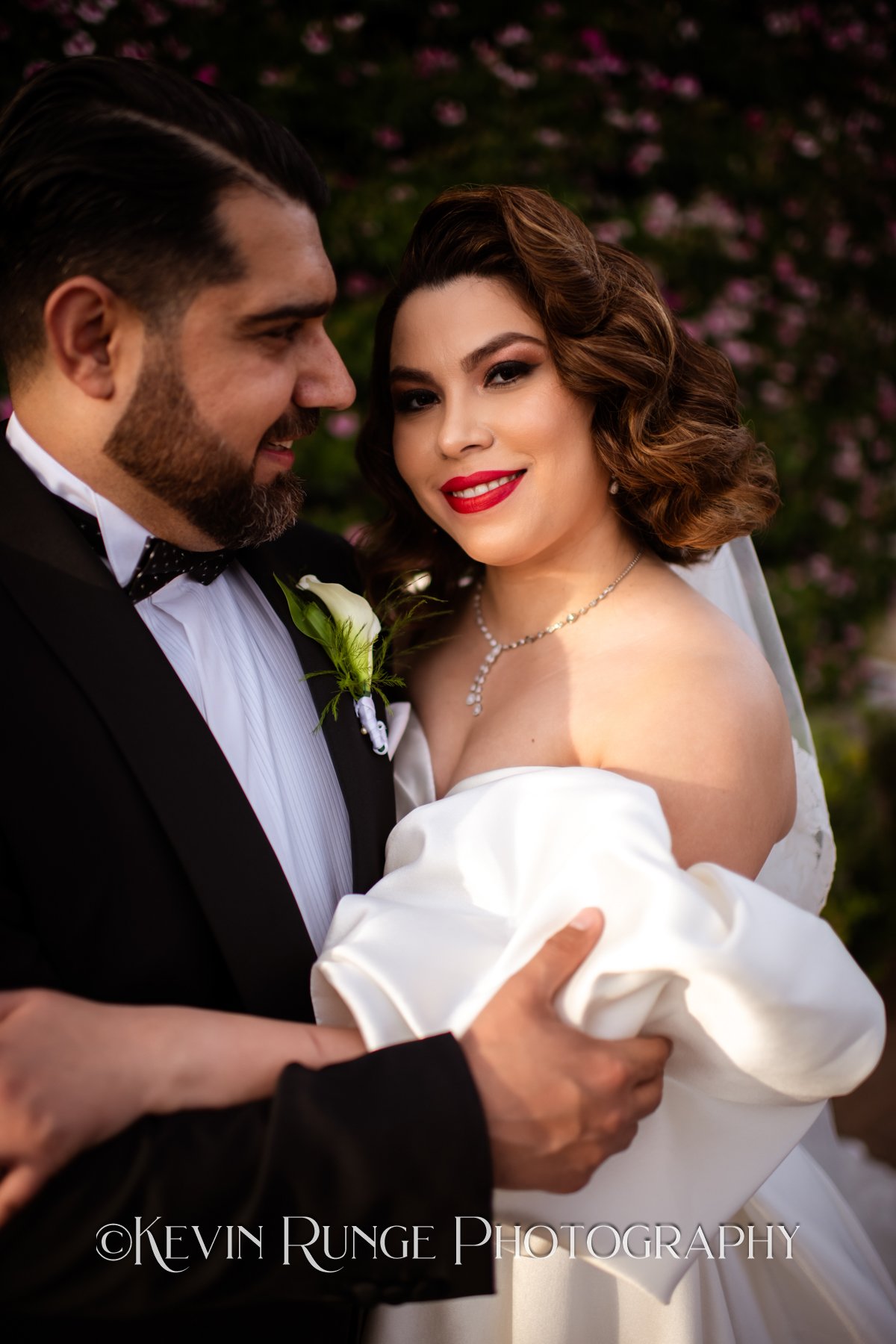 Bride and groom in wedding attire, close-up portrait outdoors with pink flowers in background, smiling and embracing.