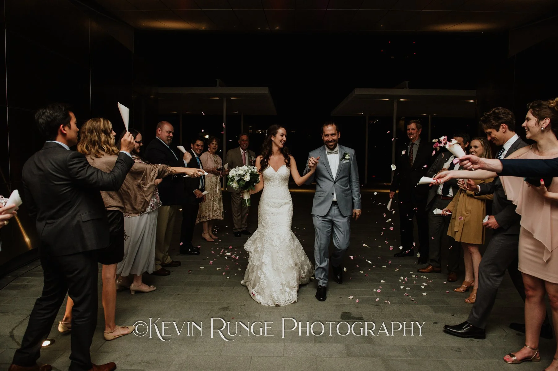 Bride and groom walking through a celebration with friends and family throwing flower petals at night.
