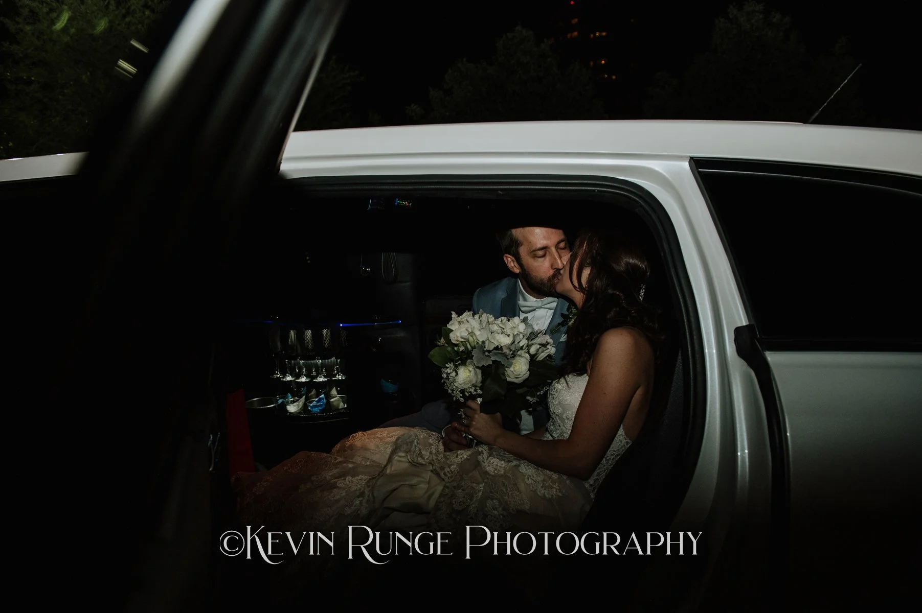 A bride and groom share a kiss inside a limousine at night, with the bride holding a bouquet of white flowers.