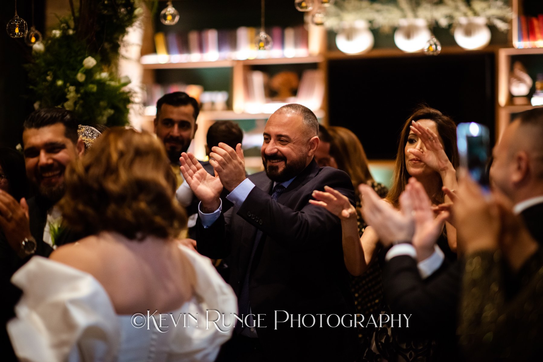 A group of people at a celebration or party, clapping and smiling, in a warmly lit room with shelves and hanging decorative ornaments, including clear baubles.