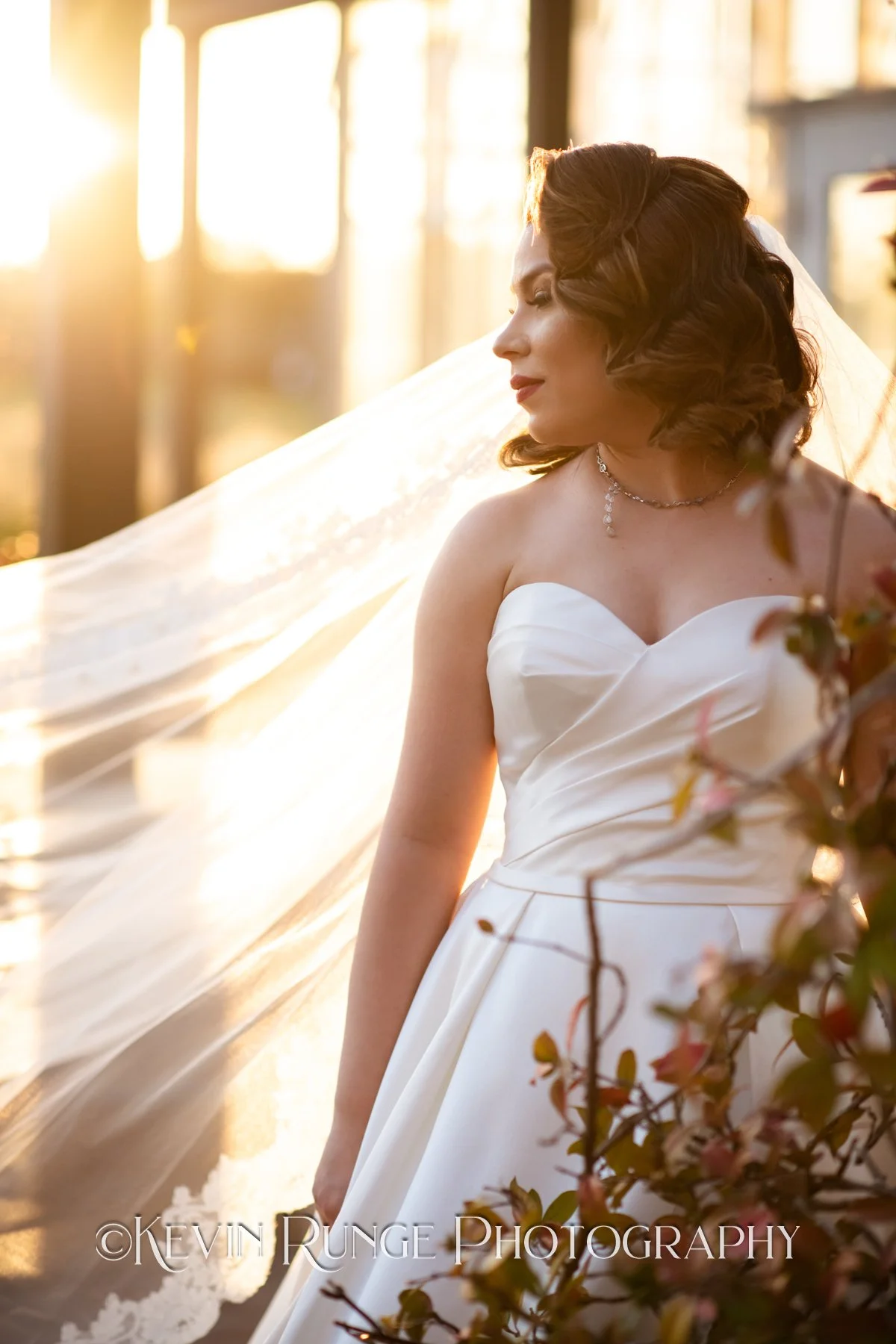 A bride stands outdoors during sunset, wearing a white strapless wedding dress with a sweetheart neckline, and a delicate necklace. Her dark hair is styled in vintage waves, and she gazes to the side with the sunlight illuminating her face and veil. 