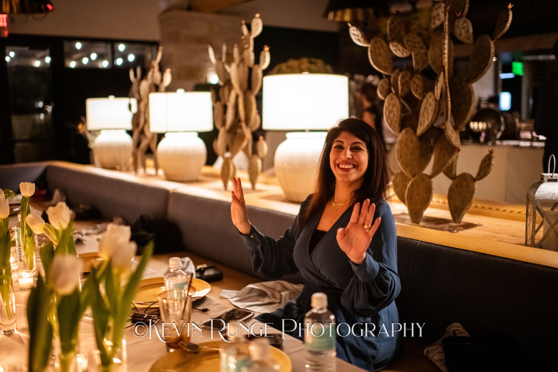 A woman with dark hair smiling and making a peace sign with her hands at a dinner table in a warmly lit restaurant.