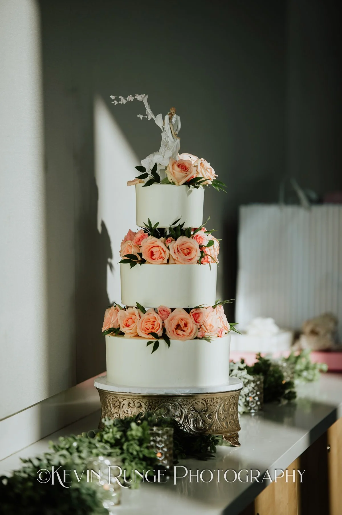 A multi-tiered wedding cake decorated with pink roses and a bride and groom cake topper, placed on an ornate silver stand.