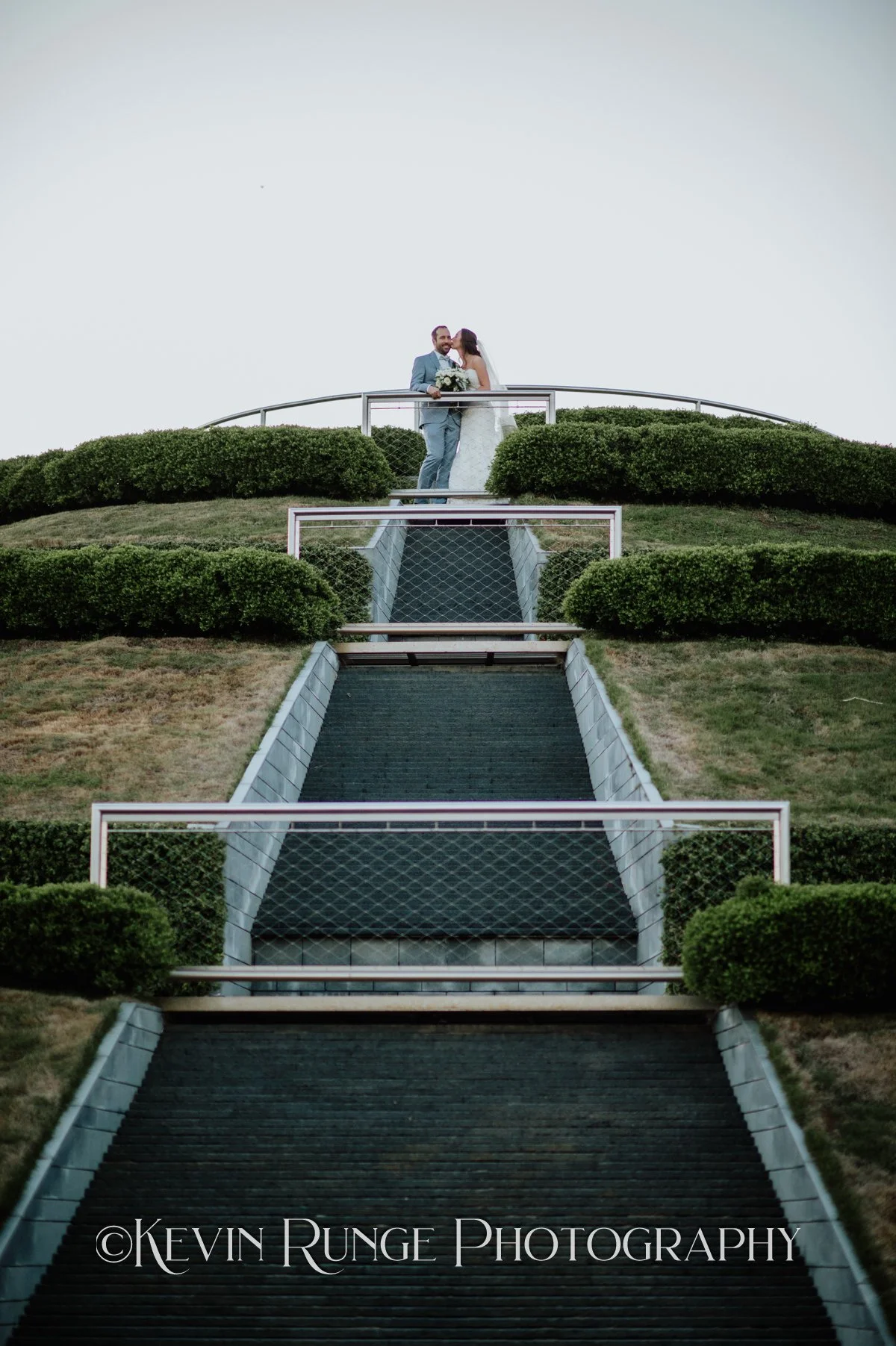 A bride and groom standing close together at the top of a set of outdoor stairs, surrounded by shrubbery, with the bride holding a bouquet of flowers.