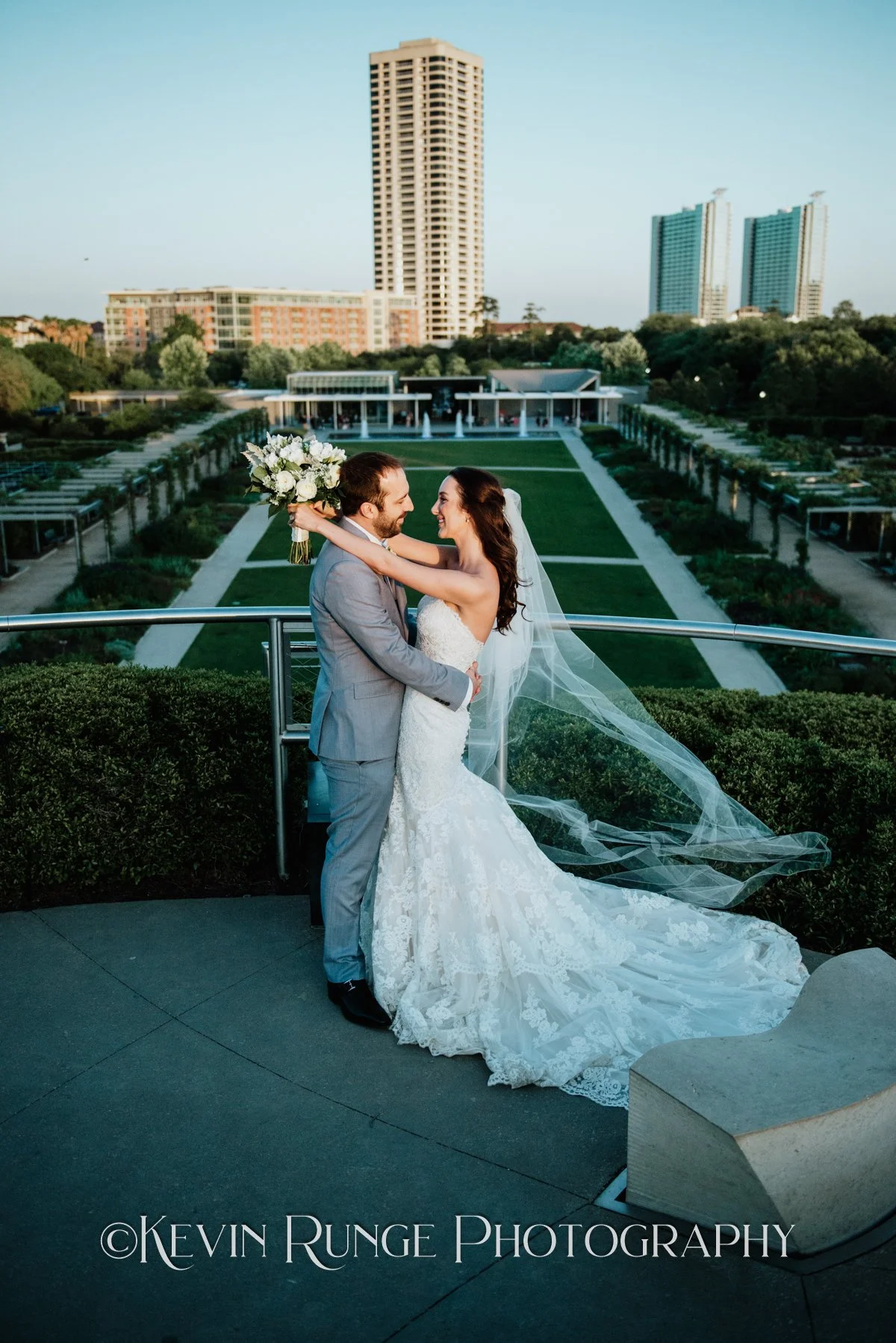 A bride and groom embrace on a balcony overlooking a cityscape with tall buildings, green trees, and a park with pathways, during sunset or early evening.