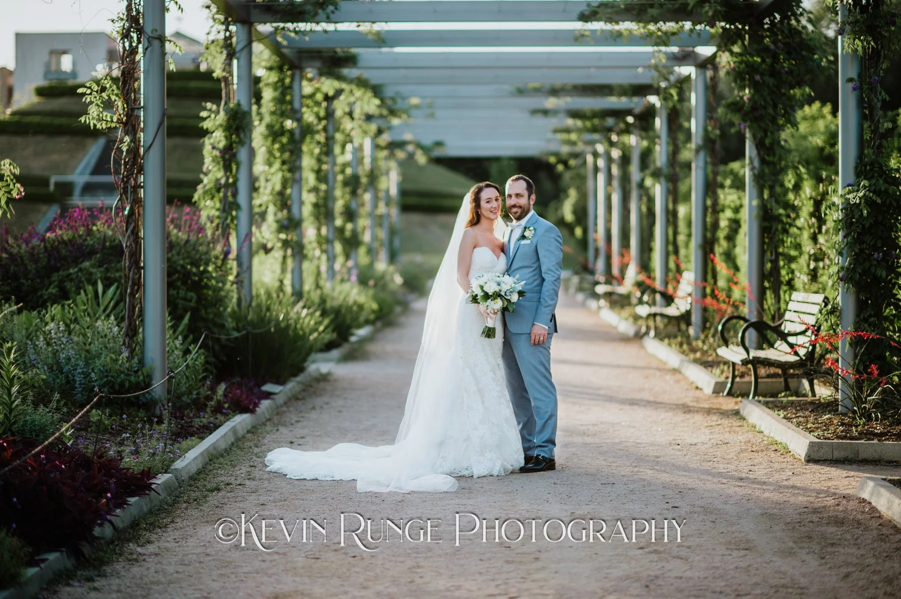 A newlywed couple standing on a garden path with greenery and flowers on both sides, under a pergola structure, during daylight. The bride is in a white wedding gown holding a bouquet, and the groom is in a light gray suit. They are smiling and posin