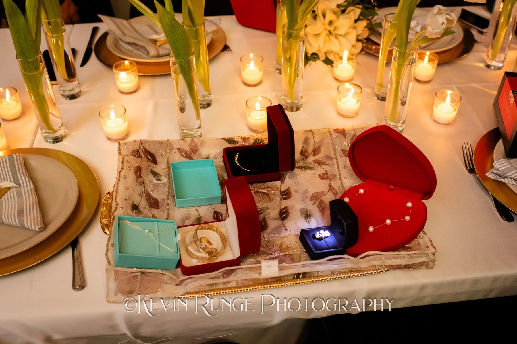 A table with lit candles, tall vases with white and yellow flowers, and a jewelry display with boxes and necklaces.