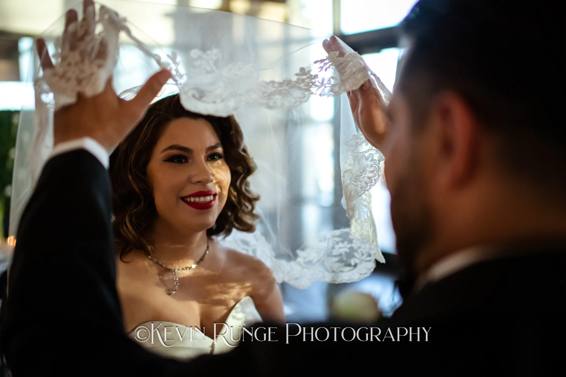 A bride with dark curly hair and red lipstick smiles as a groom lifts her veil during their wedding ceremony, with sunlight coming through the windows behind them.
