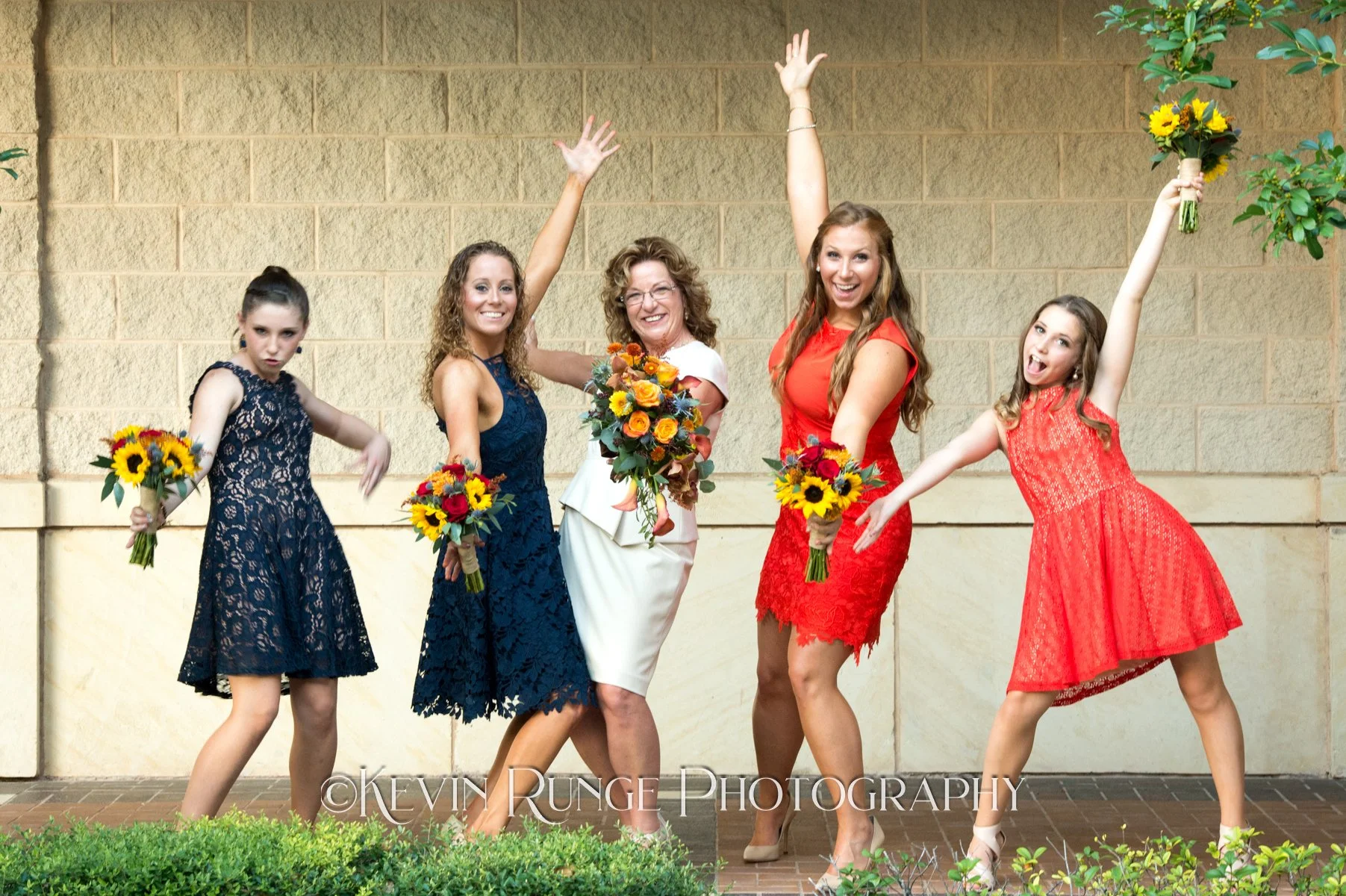 Five women, including a bride and four bridesmaids, pose happily with bouquets of flowers against a beige brick wall, celebrating a wedding.