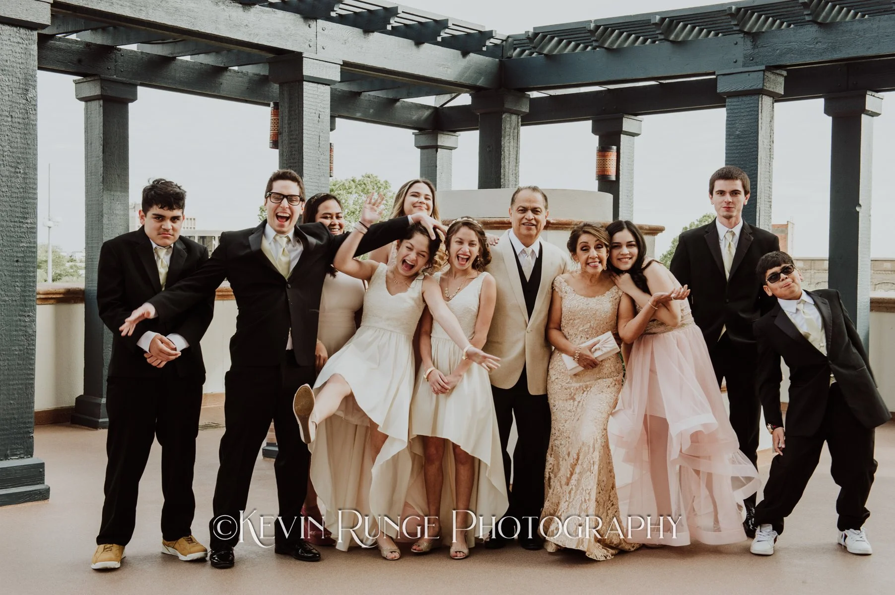 A group of people dressed in formal attire, including men in suits and women in dresses, posing together outdoors under a black wooden pergola. Some are smiling, making funny faces, or striking playful poses.
