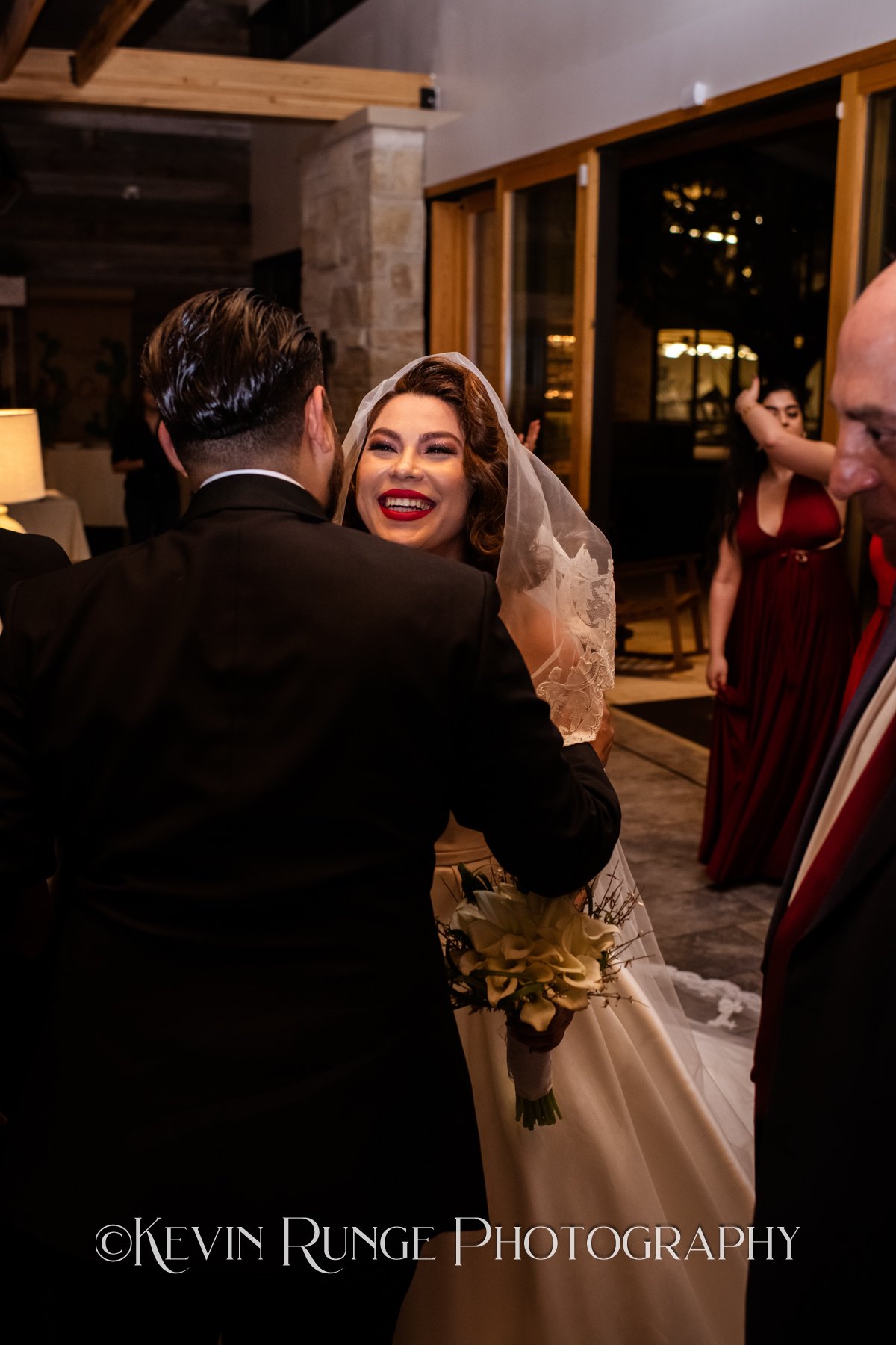 A bride with curly hair and red lipstick smiling and hugging a groom in a black suit at a wedding reception. The bride is holding a bouquet of white calla lilies. There are other guests in the background, including a woman in a red dress and a man in