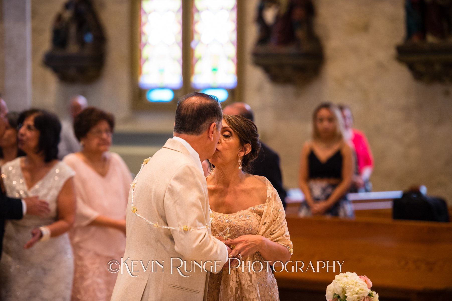 A couple sharing a kiss during a wedding ceremony in a church, with guests watching in the background.