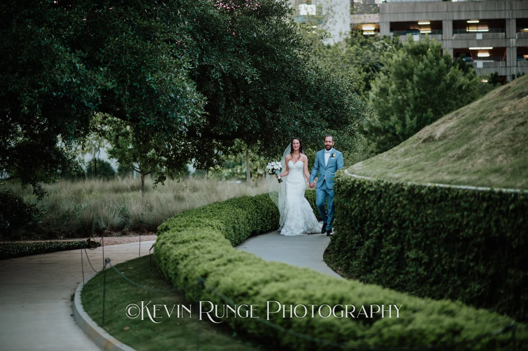 A bride and groom walking hand in hand along a curved pathway bordered by greenery, with trees and a building in the background.