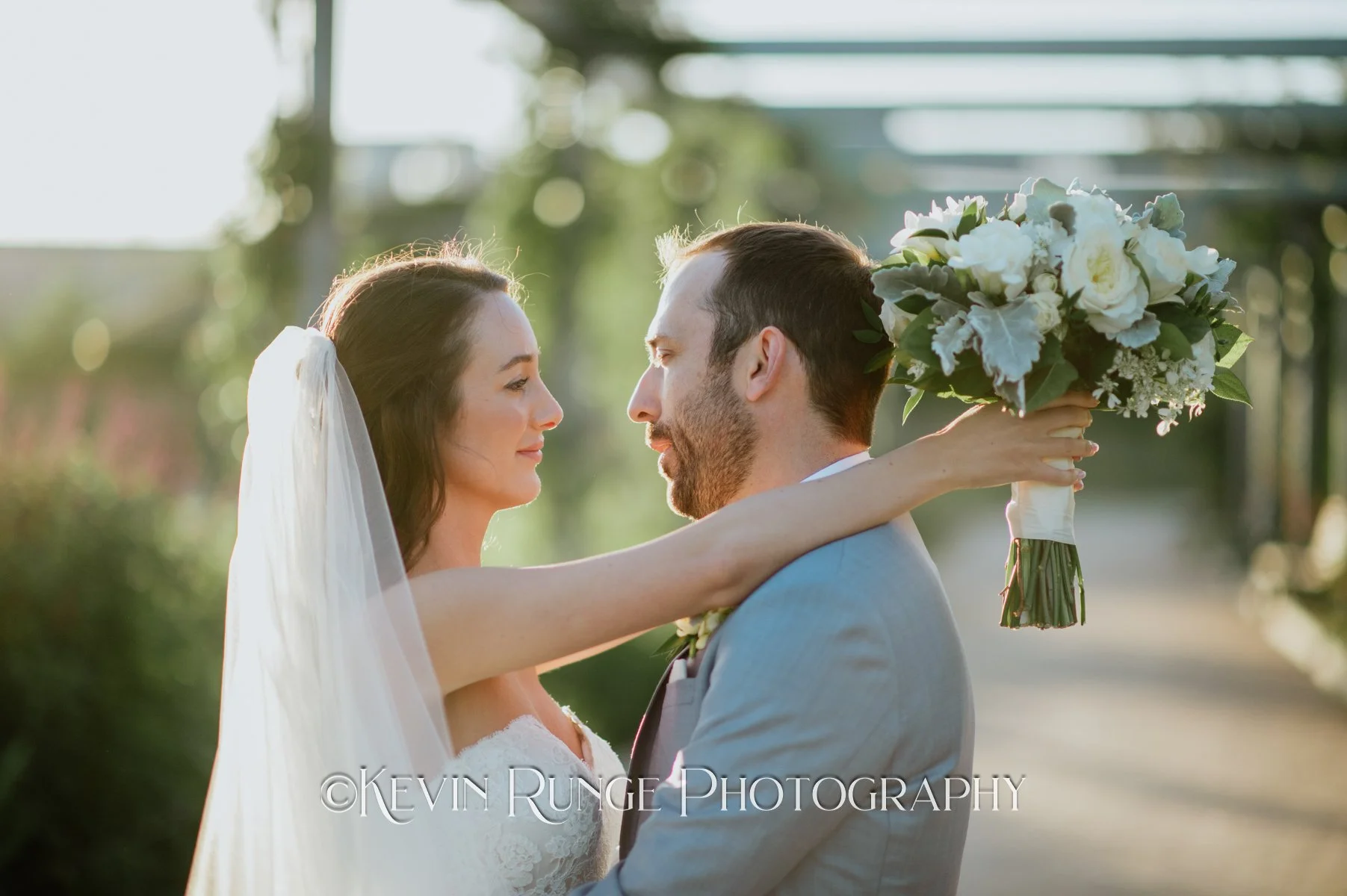 Bride and groom facing each other outdoors, the bride holding a bouquet of white flowers, both smiling, with sunlight and greenery in the background.