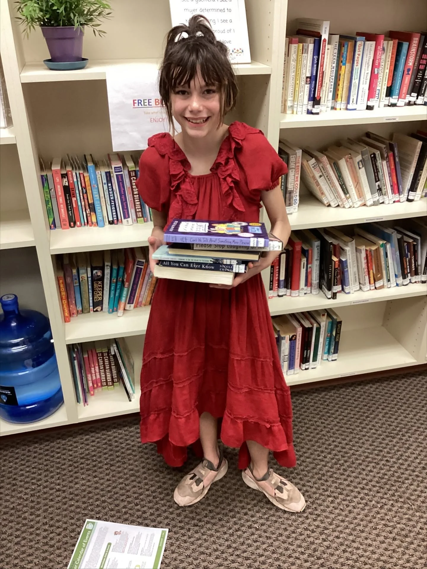 smiling little girl in red dress holds stack of books in front of a bookshelf