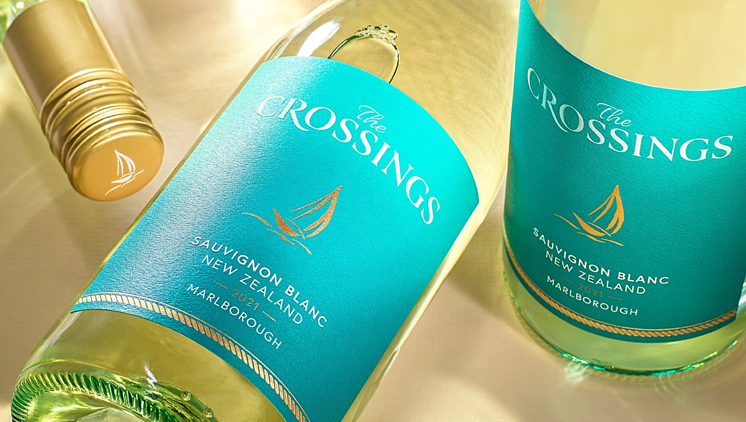 Close-up of a bottle of The Crossings Sauvignon Blanc wine from Marlborough, New Zealand, with a turquoise label and gold foil on the neck, lying on a reflective surface.