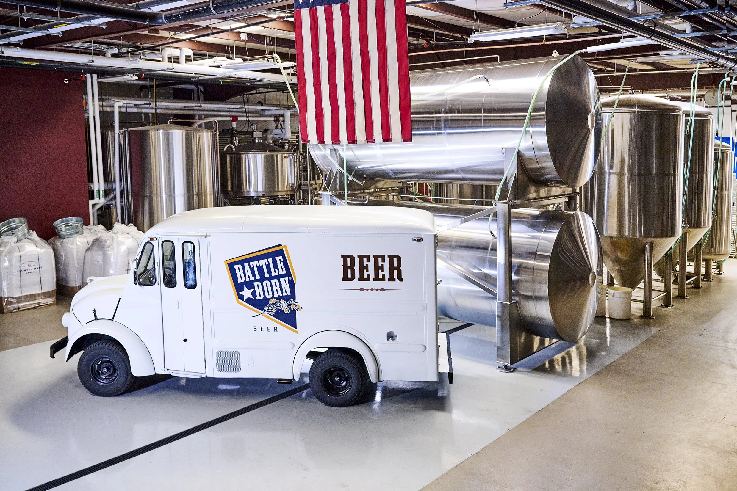 A stainless steel beer brewing tank and a beer truck with a Texas flag hanging by the ceiling in a brewery.